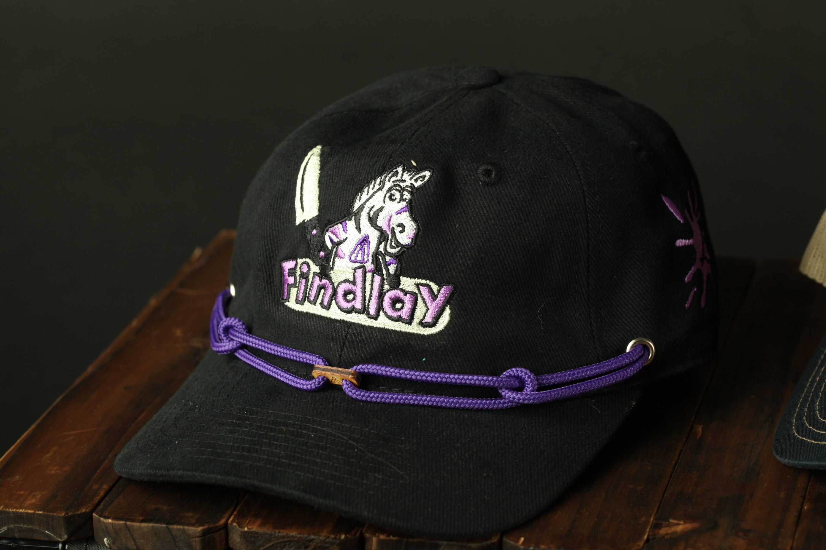 A black baseball cap with a purple and white embroidered logo and a purple rope around the brim.