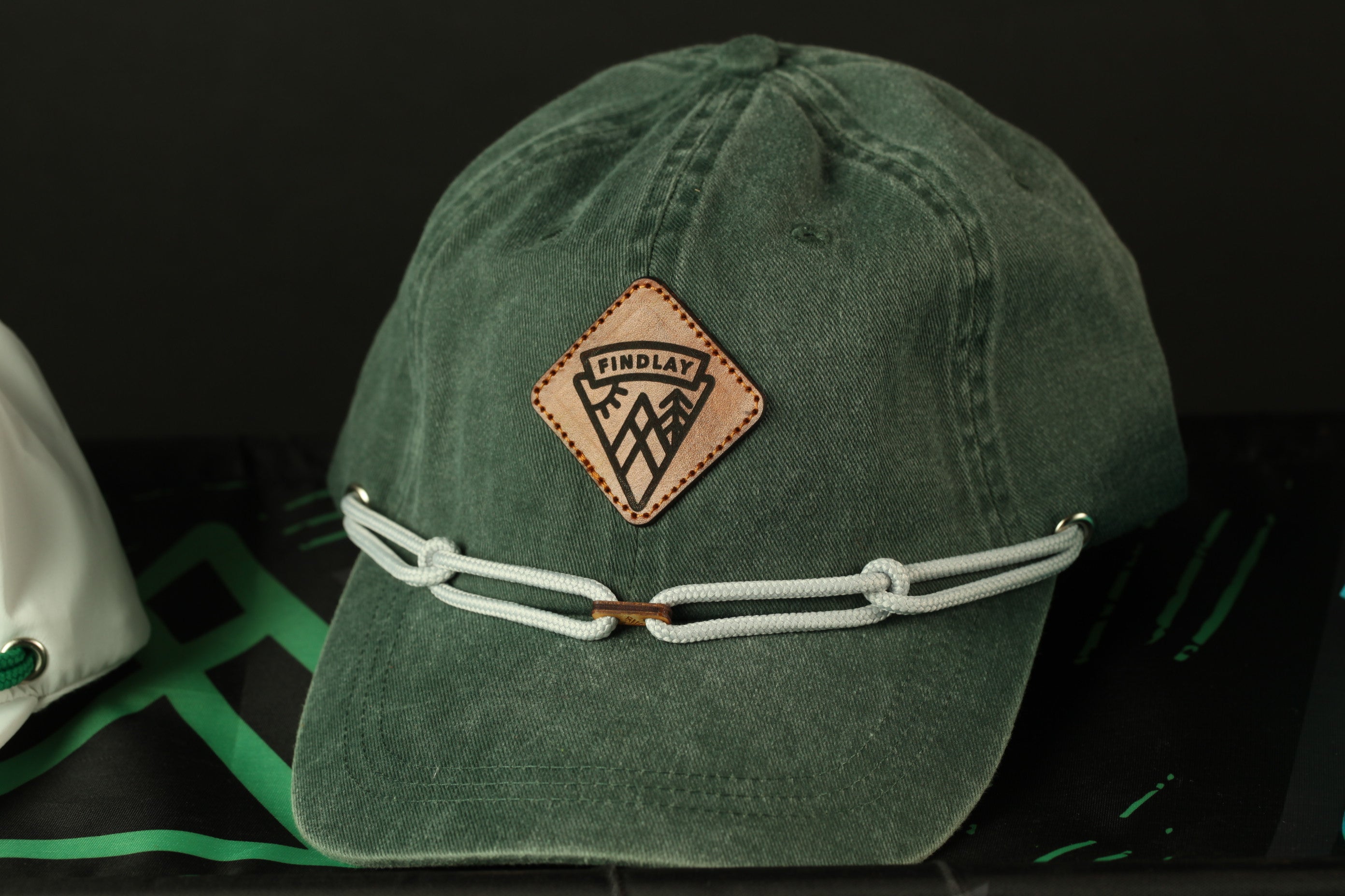 A green baseball cap with a leather patch and white braided straps is displayed against a black background.