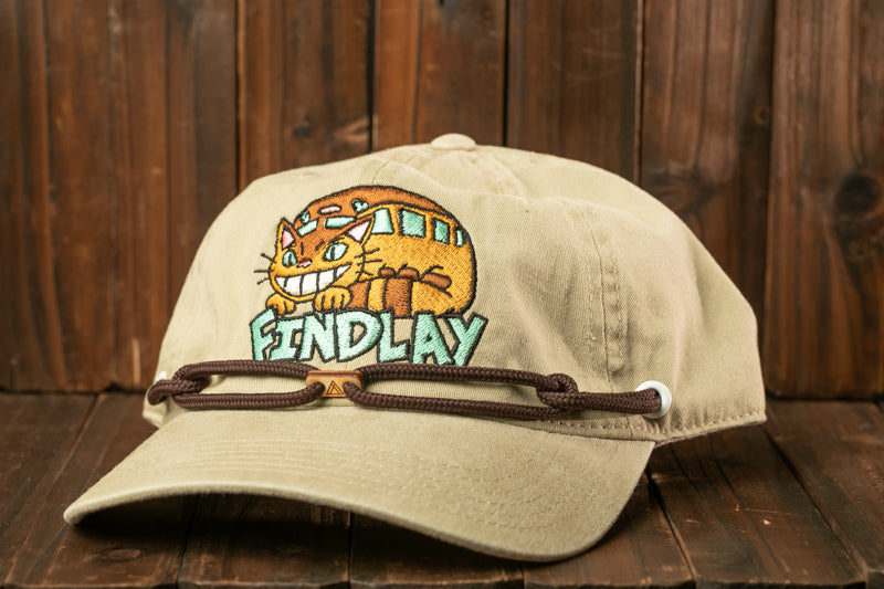 A beige baseball cap with a cartoon cat design and the text "FIND LAY" printed on it, sitting on a wooden surface.
