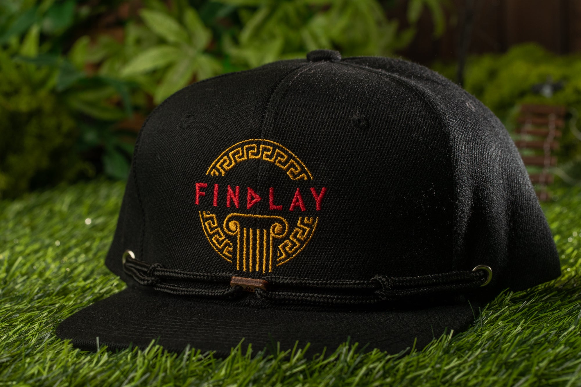 A black baseball cap with the word "FIND LAY" embroidered on it, sitting on a grassy surface.