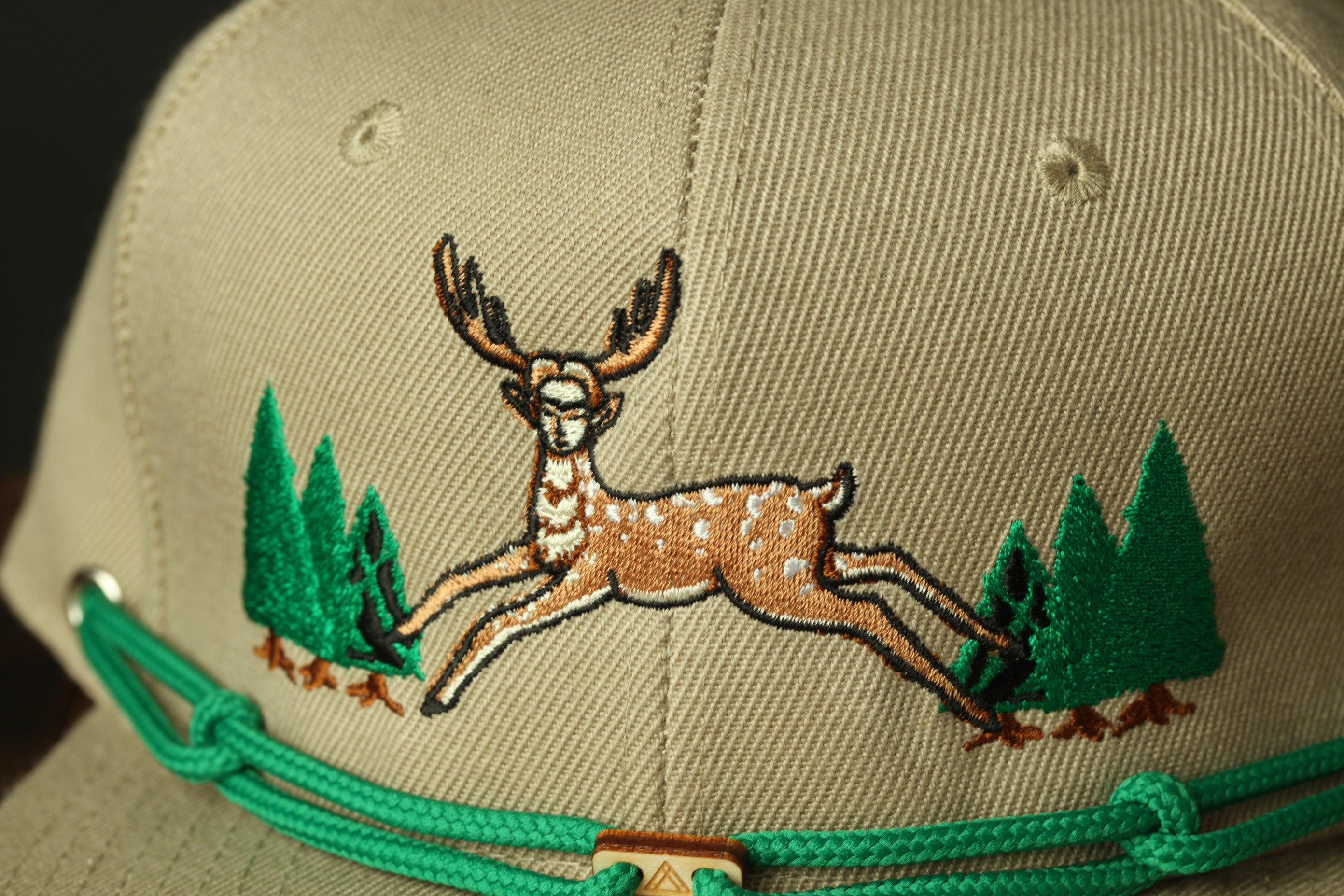 A close-up view of a beige baseball cap with a detailed embroidered design of a deer in mid-leap, surrounded by green trees.