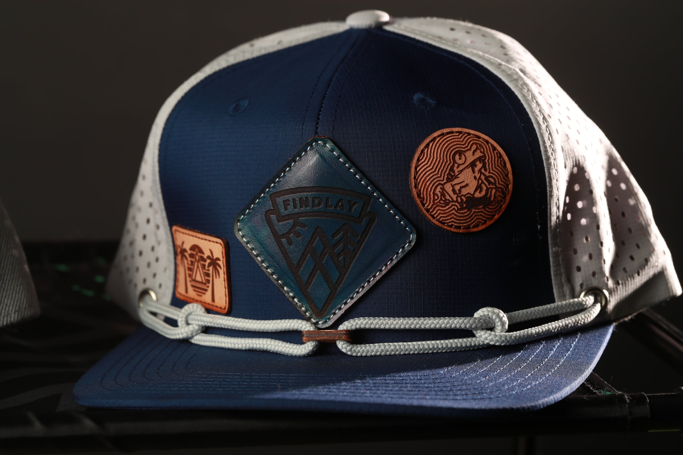 A blue baseball cap with a white perforated brim and a leather patch on the front, featuring a logo and a wave-like design.