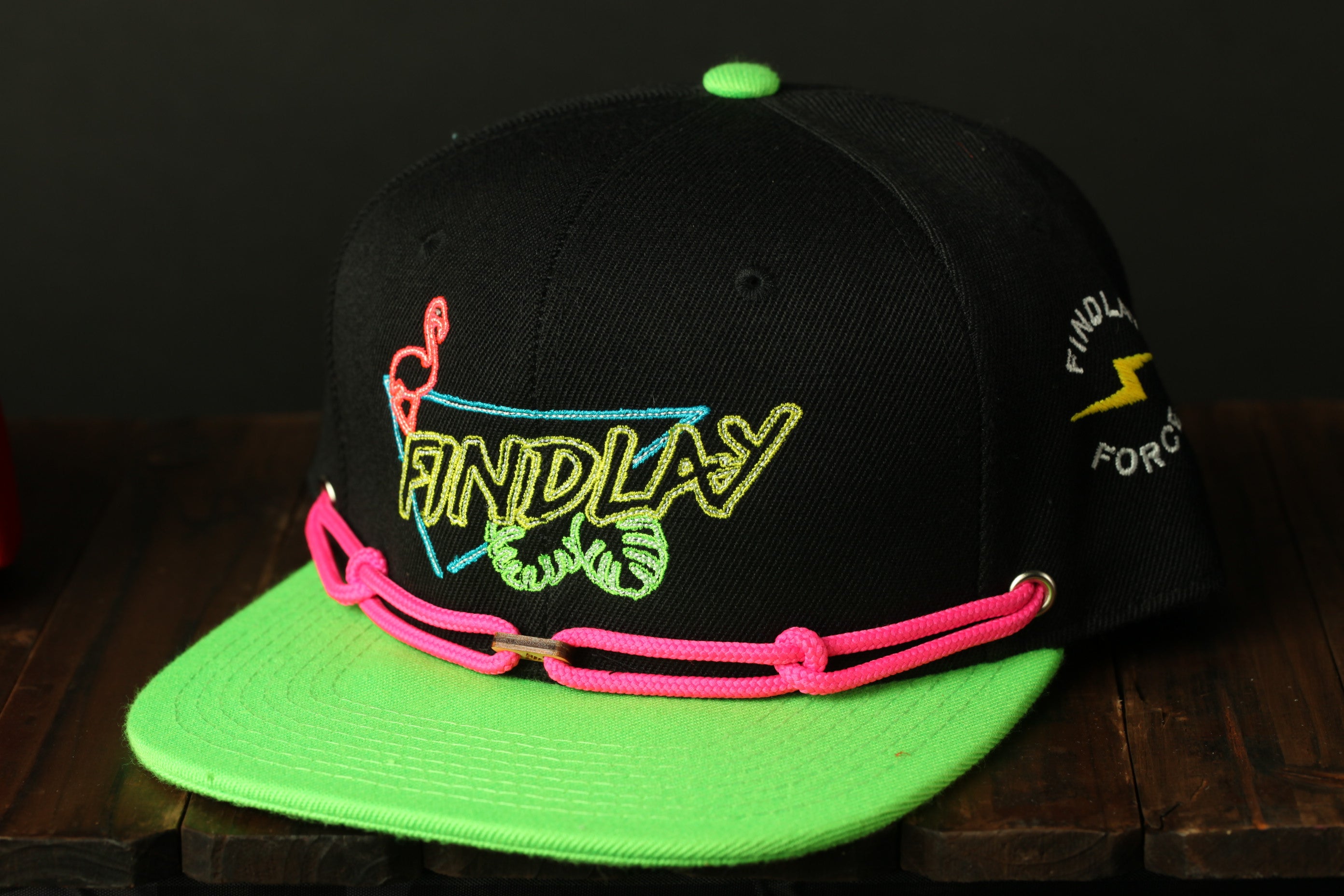 A black baseball cap with a neon green brim and a pink braided strap, featuring colorful embroidered text and graphics.