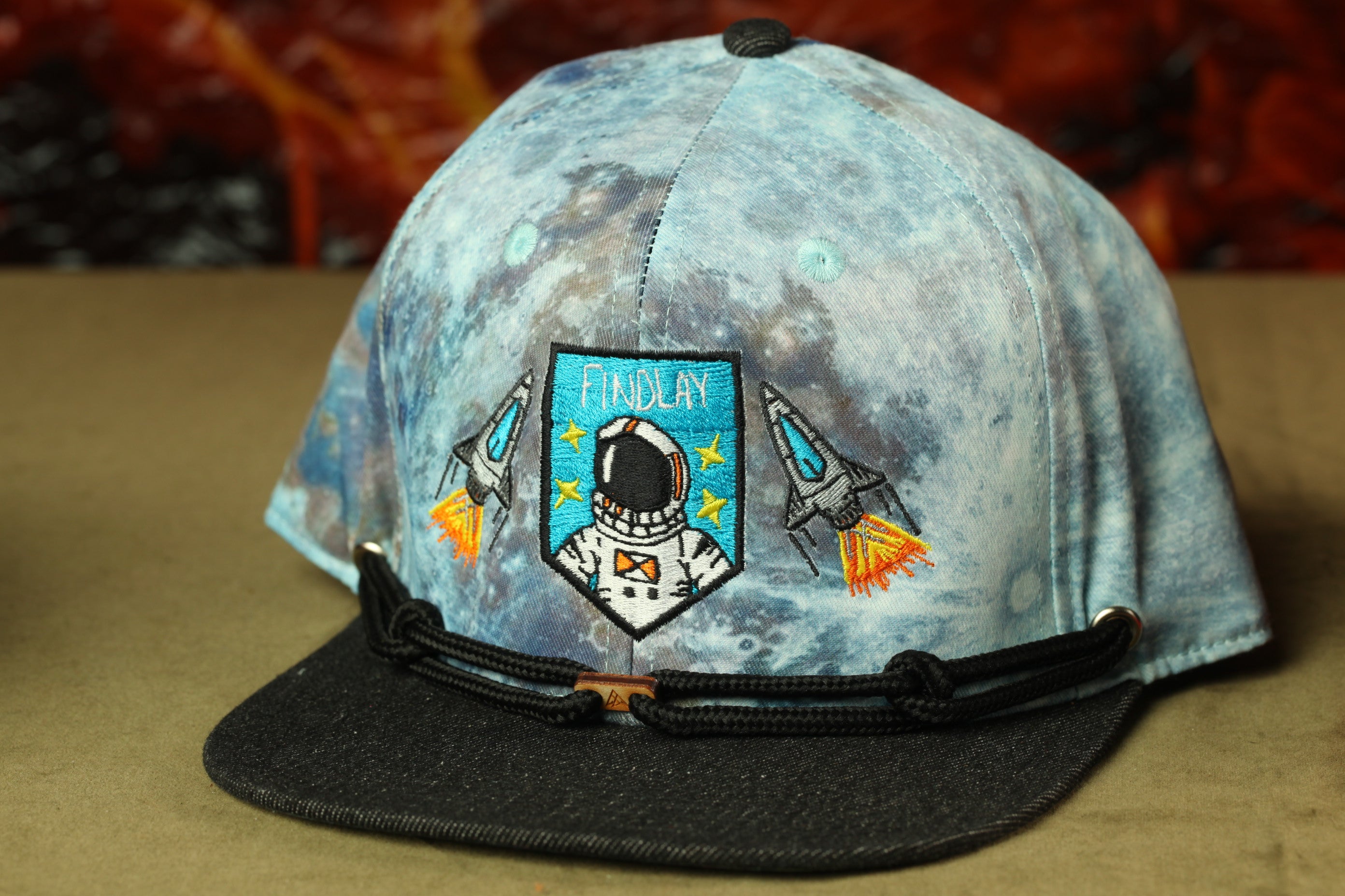 A blue baseball cap with a space-themed design, including a space suit and rocket ship motifs.