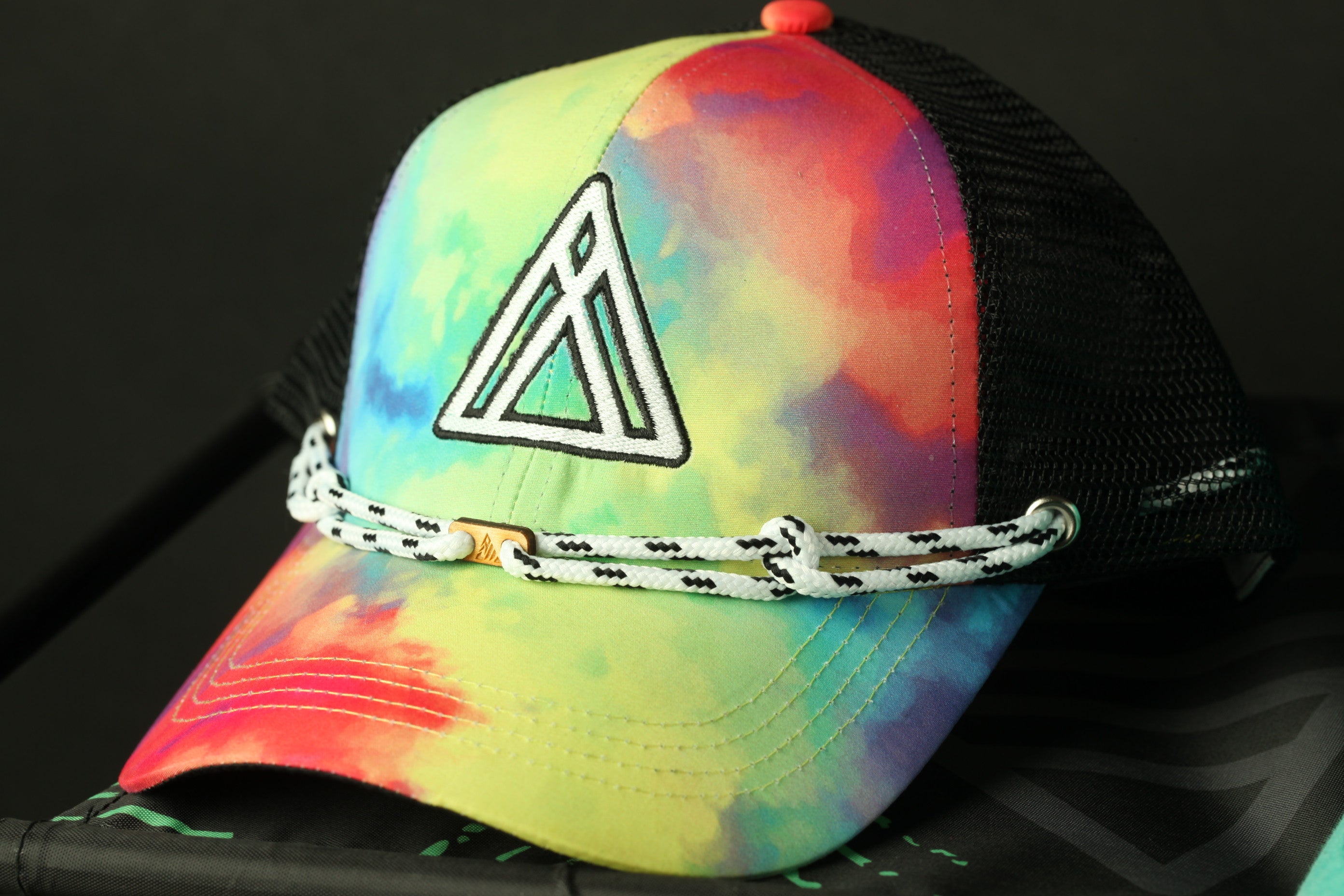 A colorful tie-dye baseball cap with a black mesh panel and a white braided rope strap.