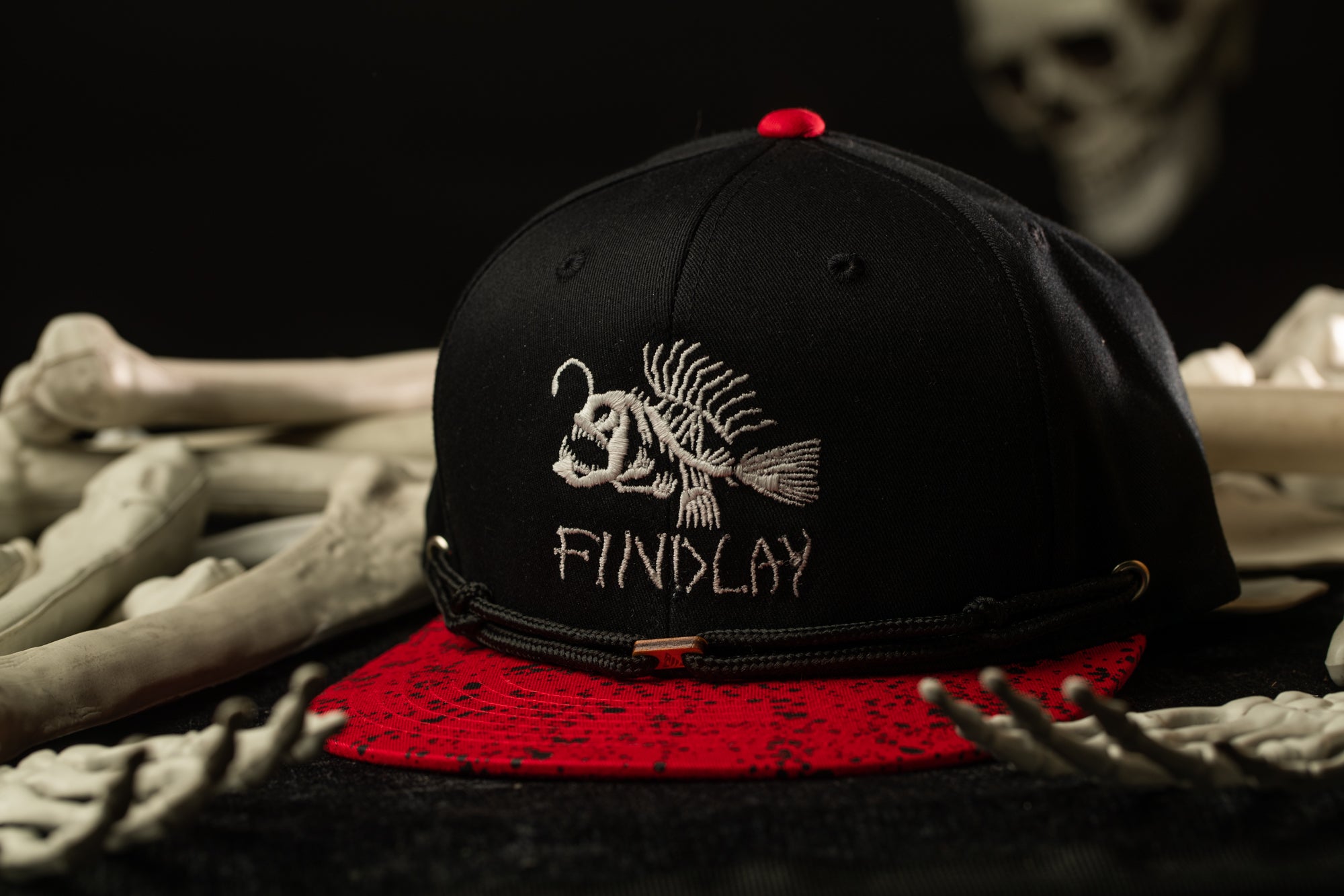A black baseball cap with a red brim and a white fish embroidered on it, placed on a red and black speckled surface.