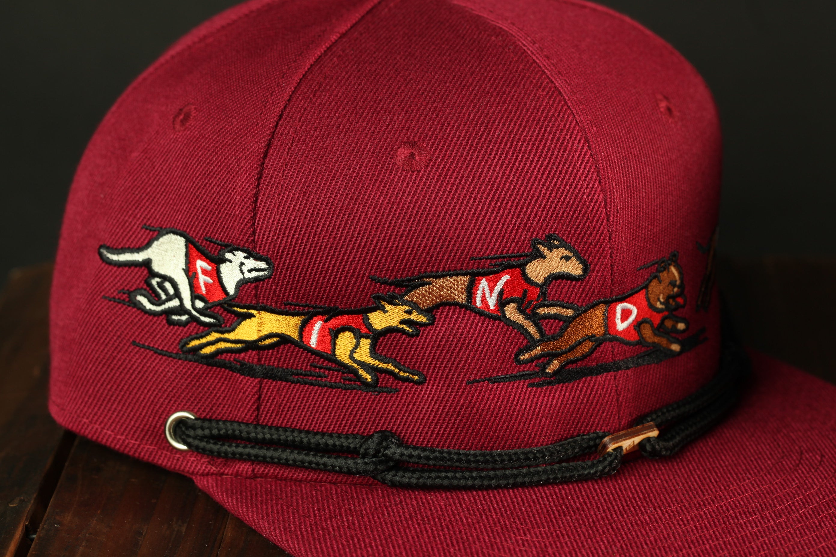 A close-up image of a red baseball cap with a black strap, featuring a design of three dogs running together.