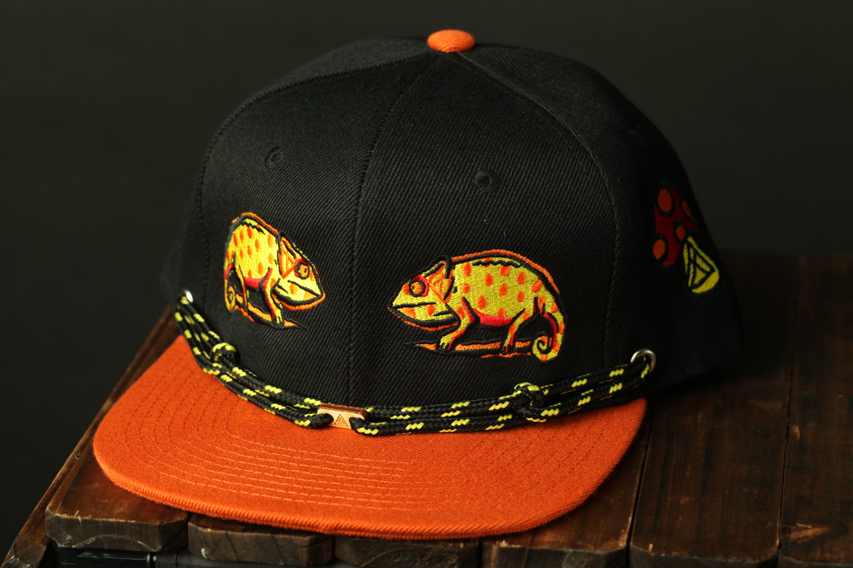 A black baseball cap with orange trim and a chameleon design on the front.