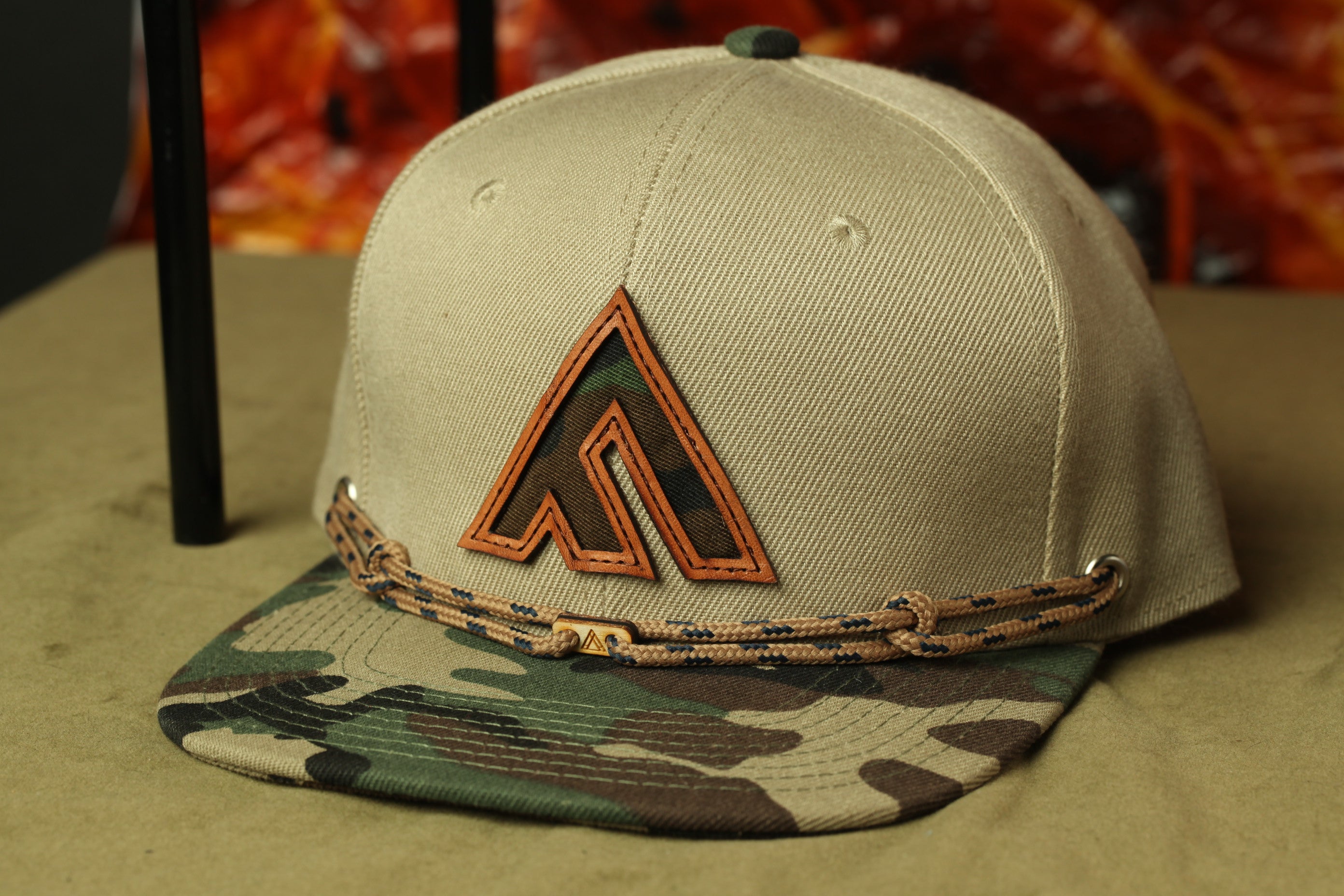 A beige baseball cap with a camouflage pattern and a logo on the front, placed on a surface with a blurred background.