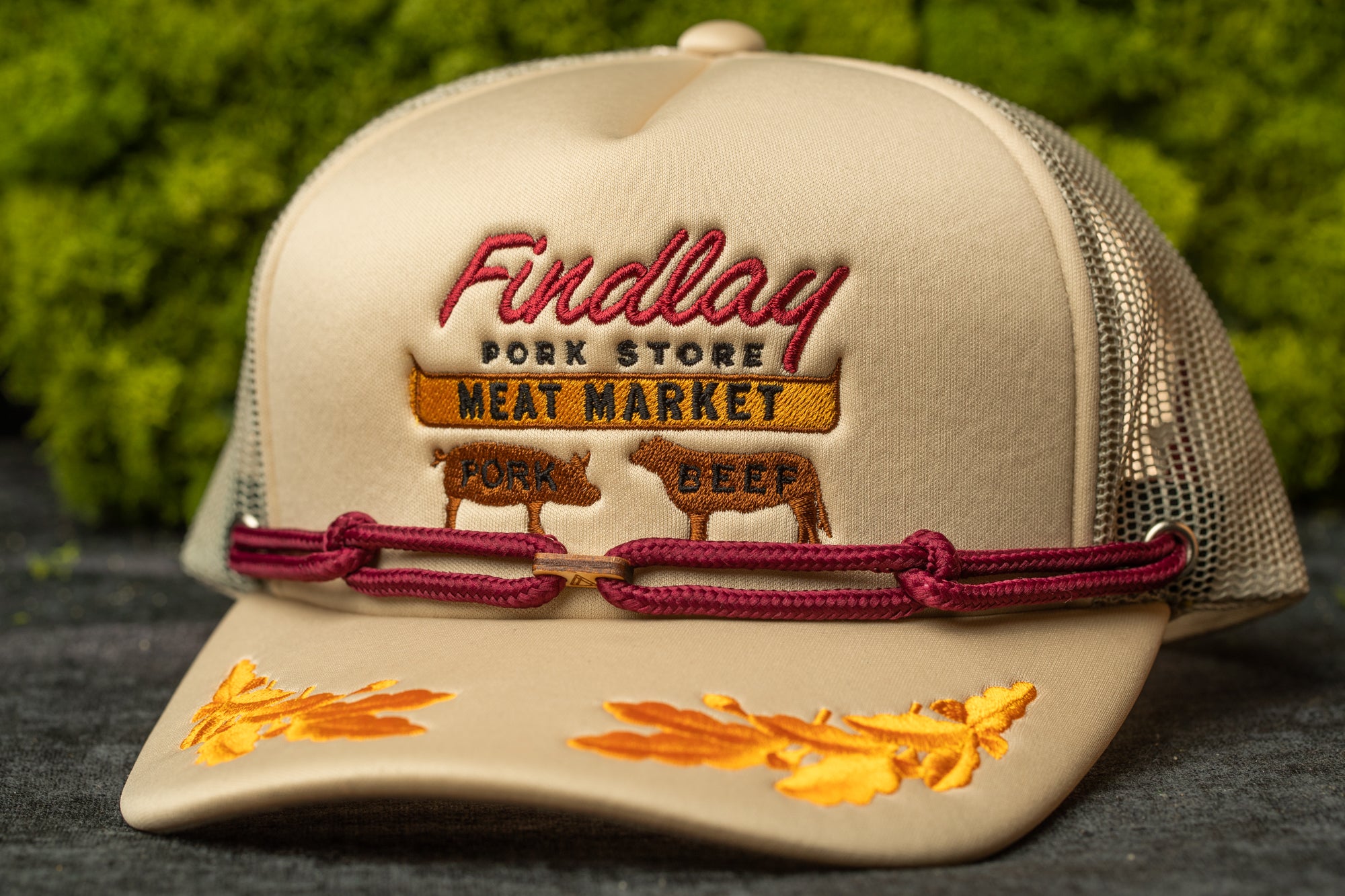 A beige baseball cap with embroidered text and images, including a pig and a cow, against a green background.