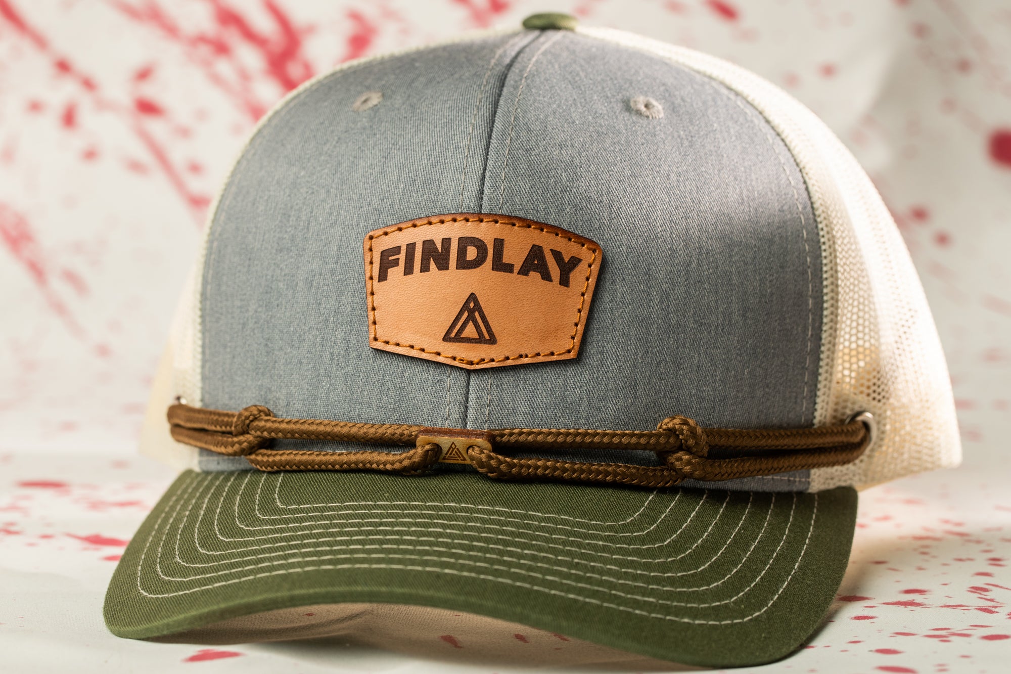 A gray and green baseball cap with a leather patch that says "FINDLAY" and a brown rope strap.