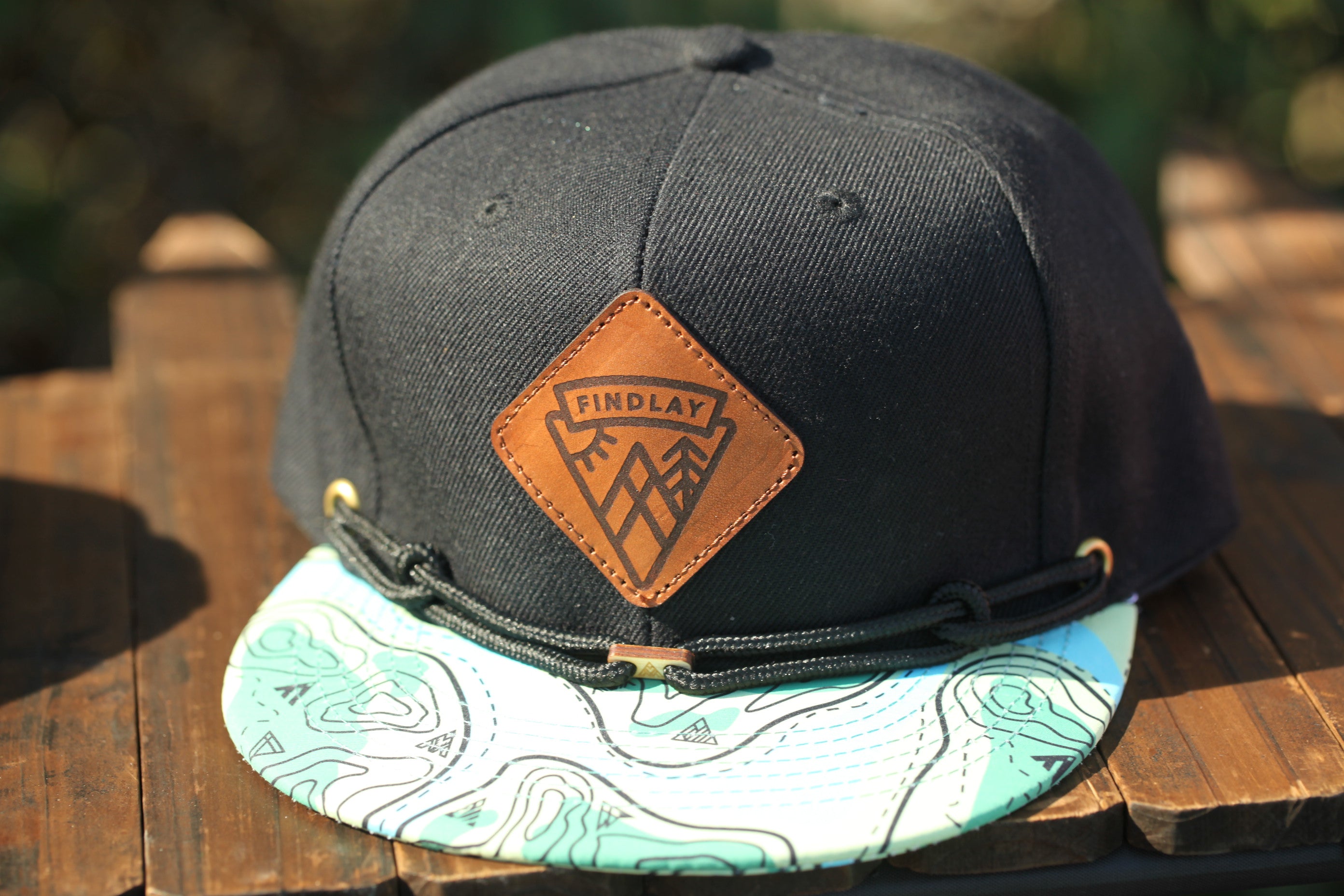 A black baseball cap with a leather patch on the front, featuring a map pattern and the word "FINDLAY" printed on it.