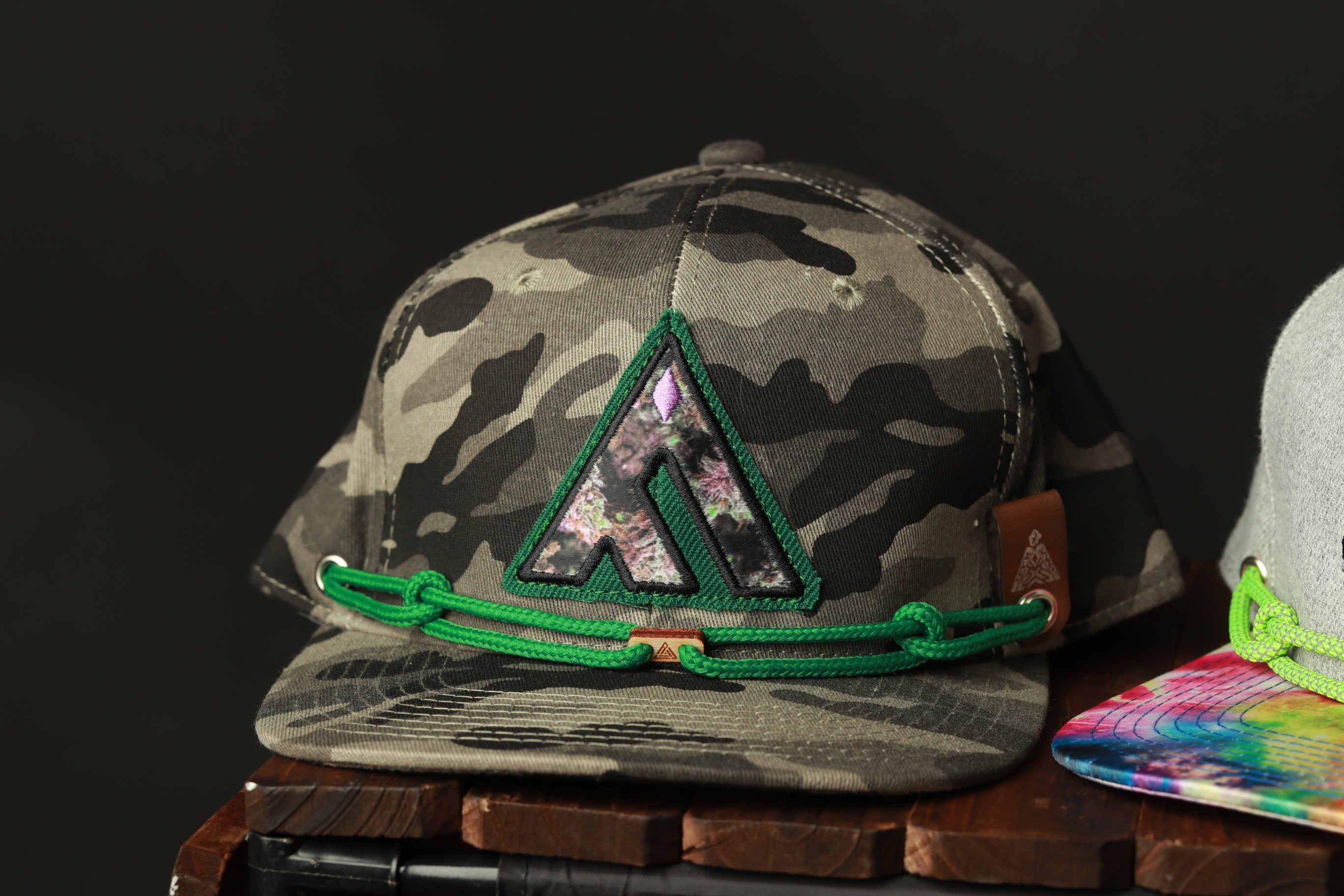 A camouflage baseball cap with a green rope strap and a unique logo on the front, displayed on a wooden surface.