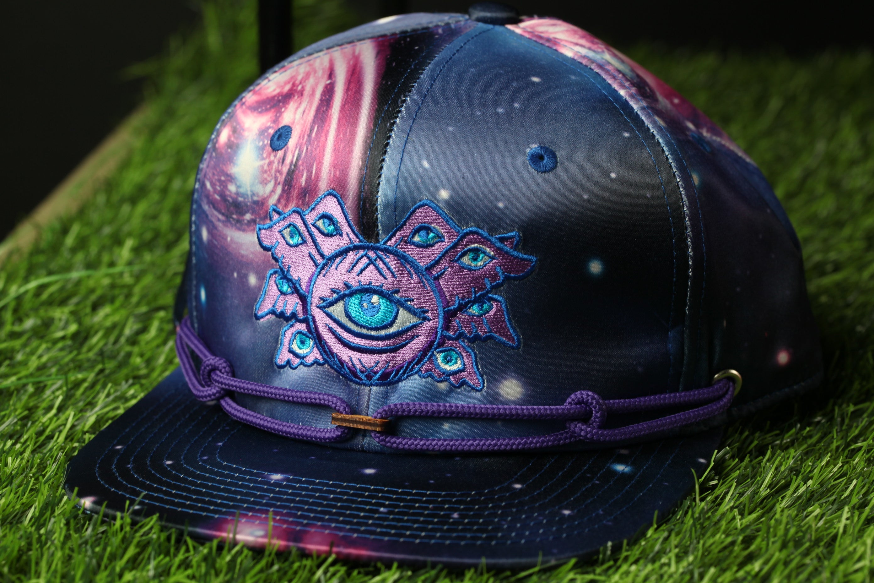 A colorful, patterned baseball cap with a large eye design on the front, sitting on a grassy surface.