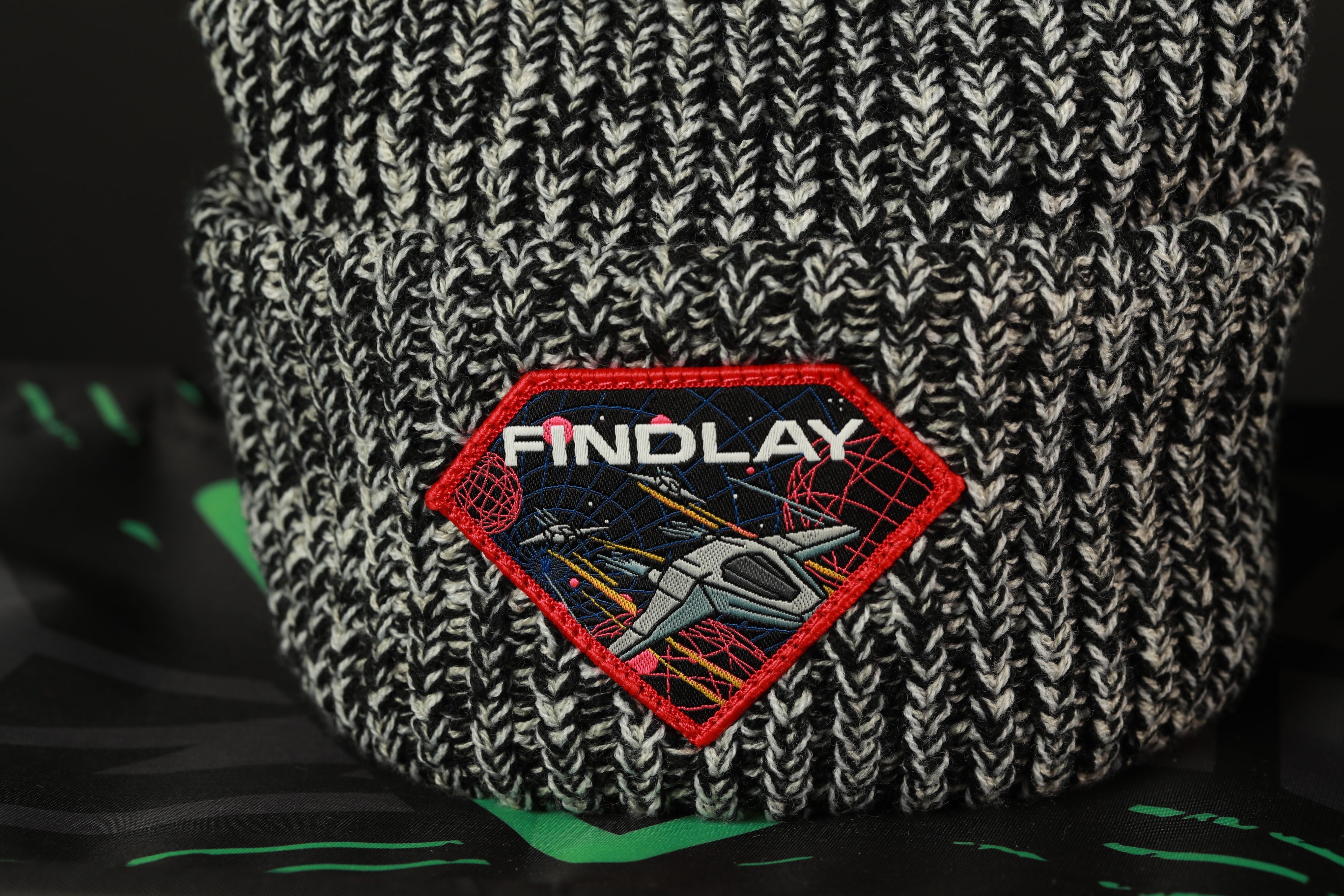 A close-up image of a knitted beanie with a red and black patch featuring a space-themed design and the word "FINDLAY" printed on it.