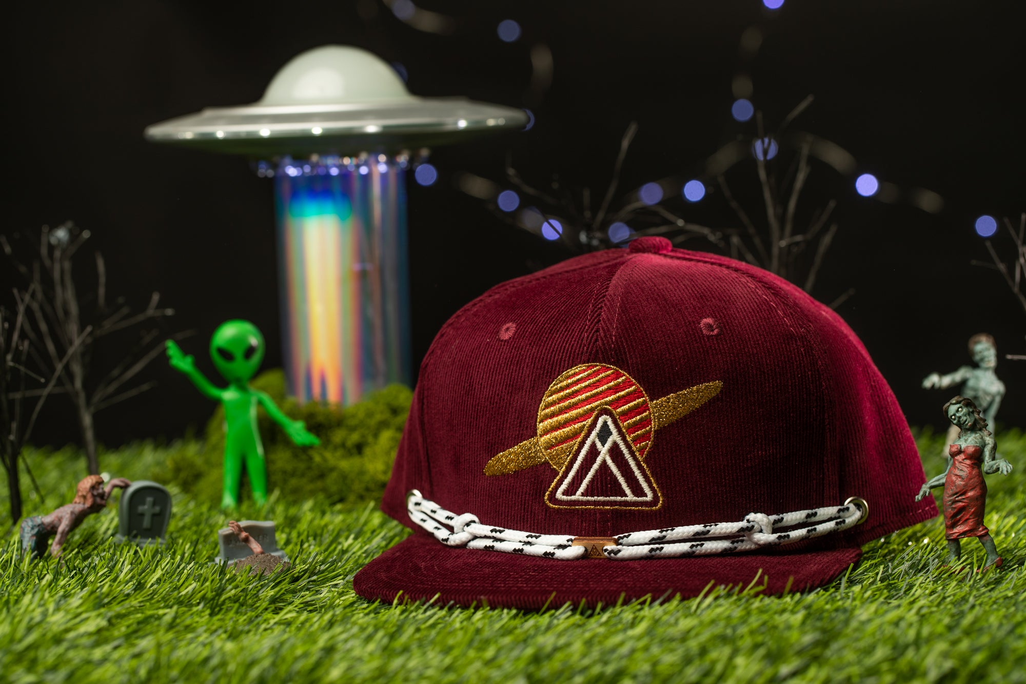 a maroon baseball cap with a sun-like emblem, surrounded by various small figures and objects, including a green alien figure, a toy spaceship, and a skull and crossbones.
