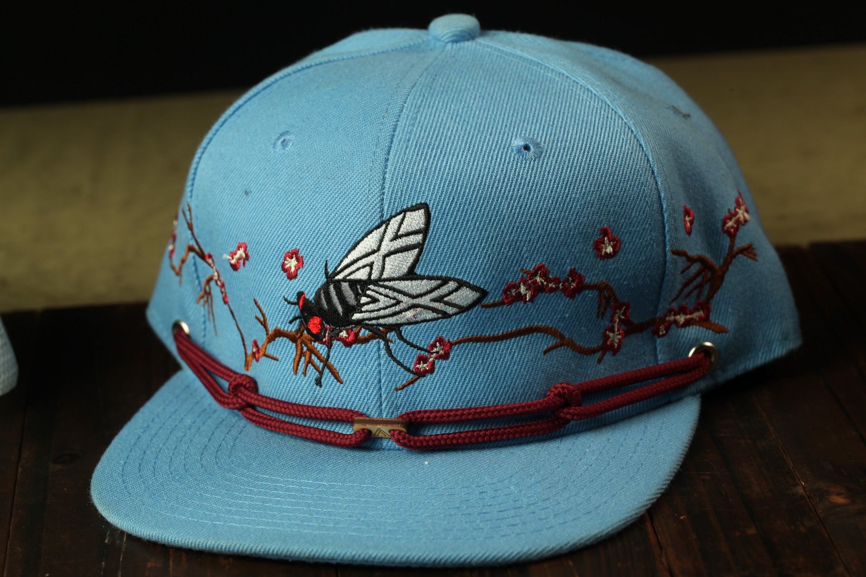 A blue baseball cap with a floral design and a butterfly motif, featuring red strings and a red buckle.