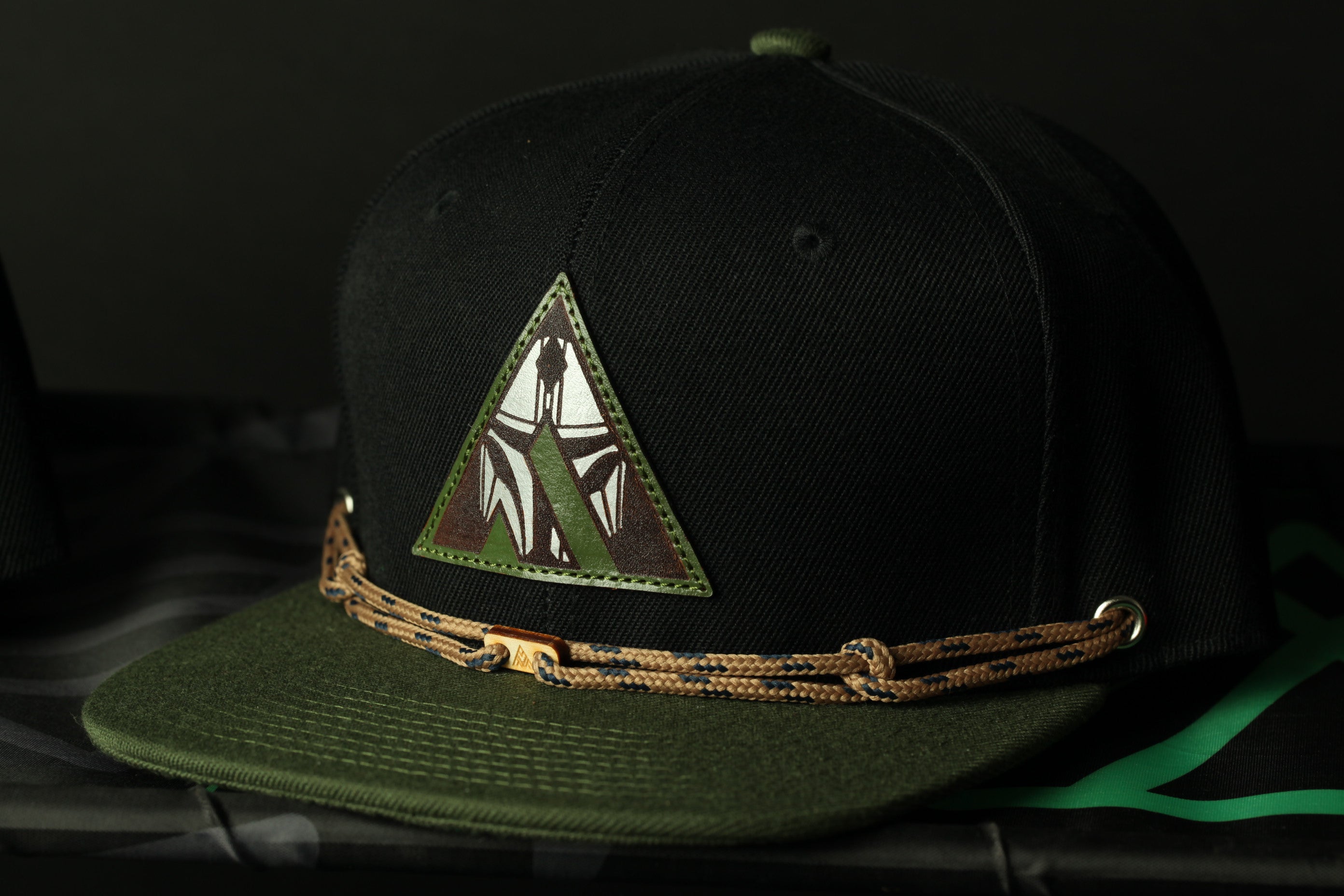 A black baseball cap with a green and brown patterned band, featuring a triangular patch with a graphic design on the front.