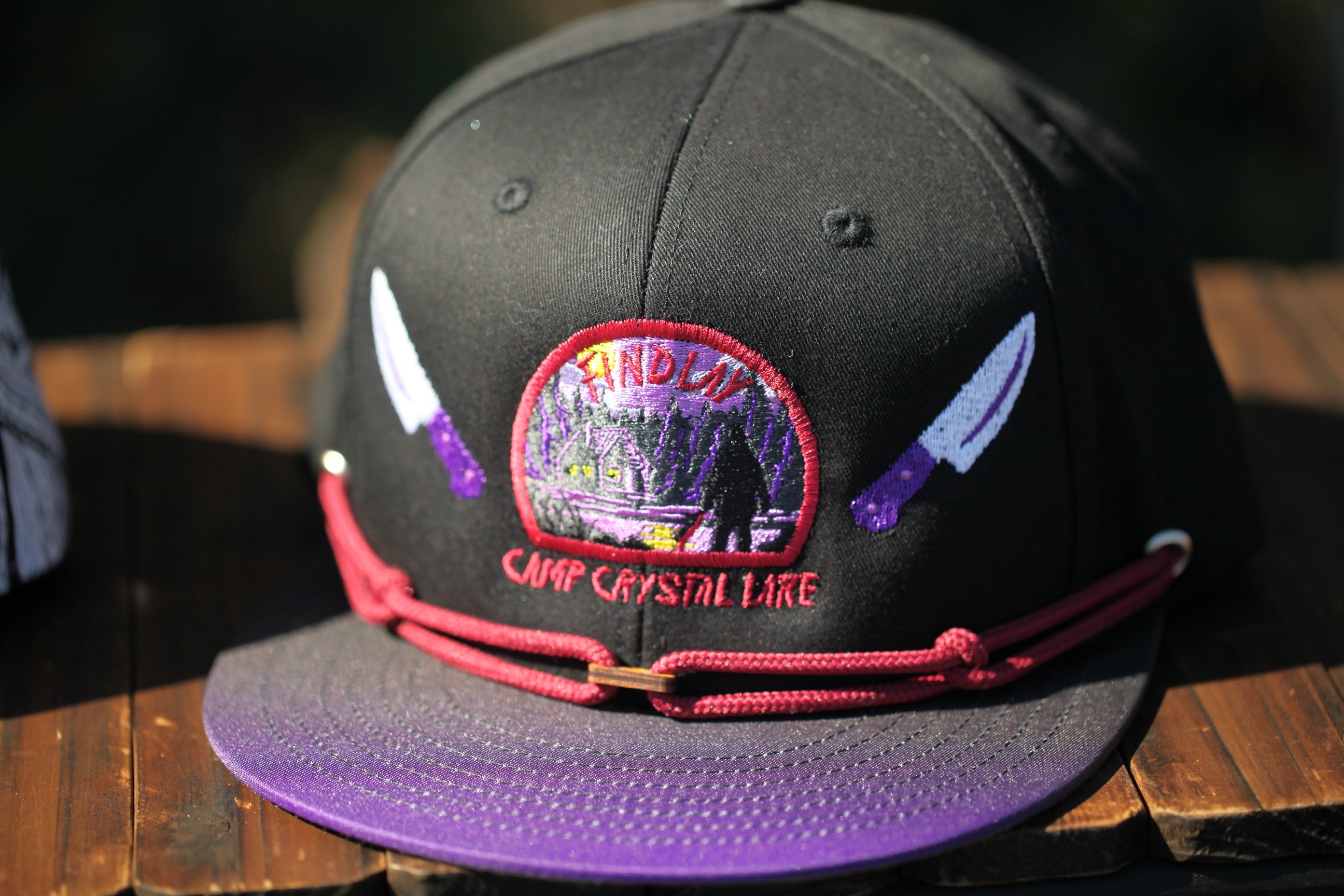 A black baseball cap with a purple brim and a red string, featuring an embroidered patch with a purple and white design and the text "Camp Crystal Lake".