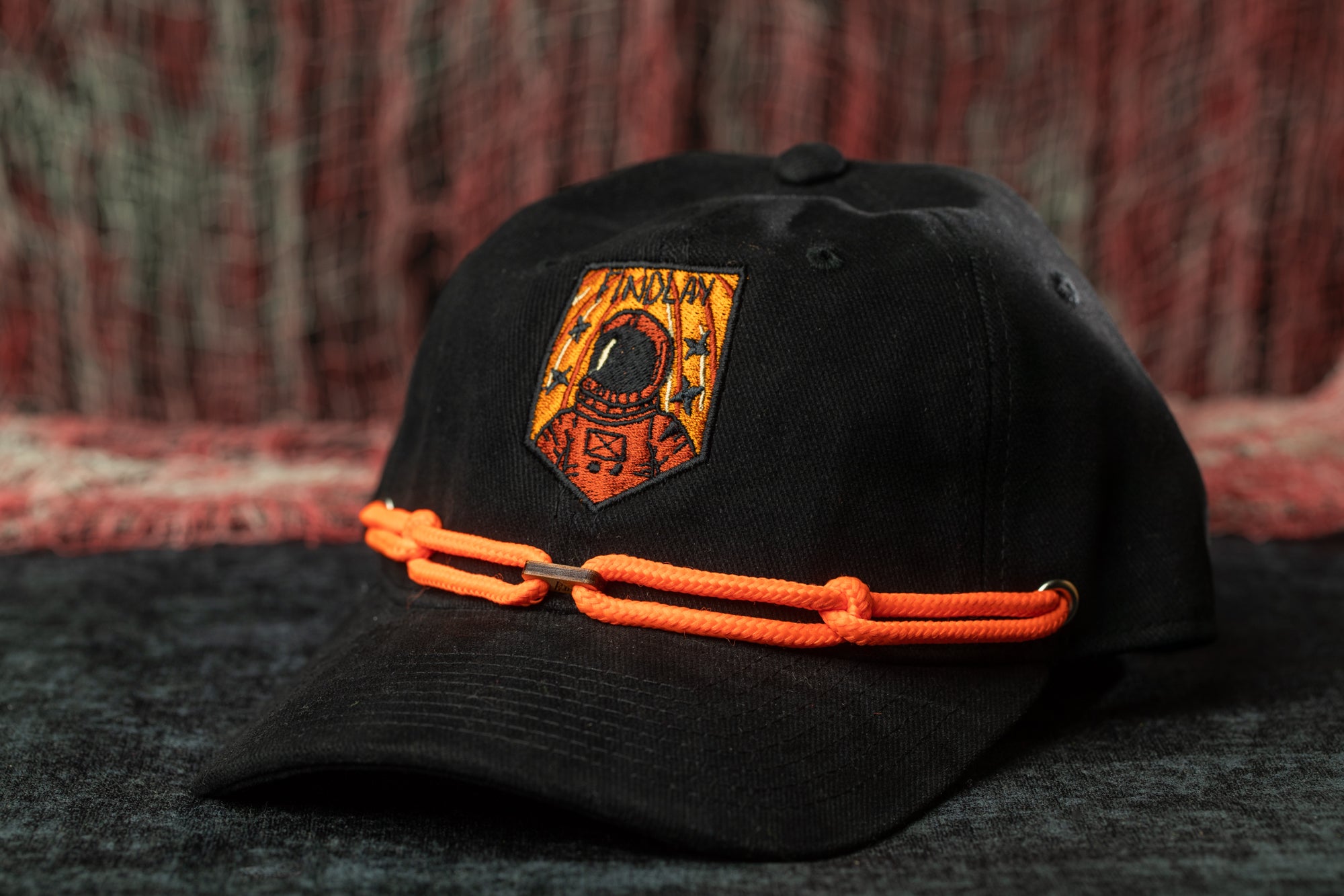A black baseball cap with an orange chain strap and an embroidered patch on the front, featuring an astronaut.