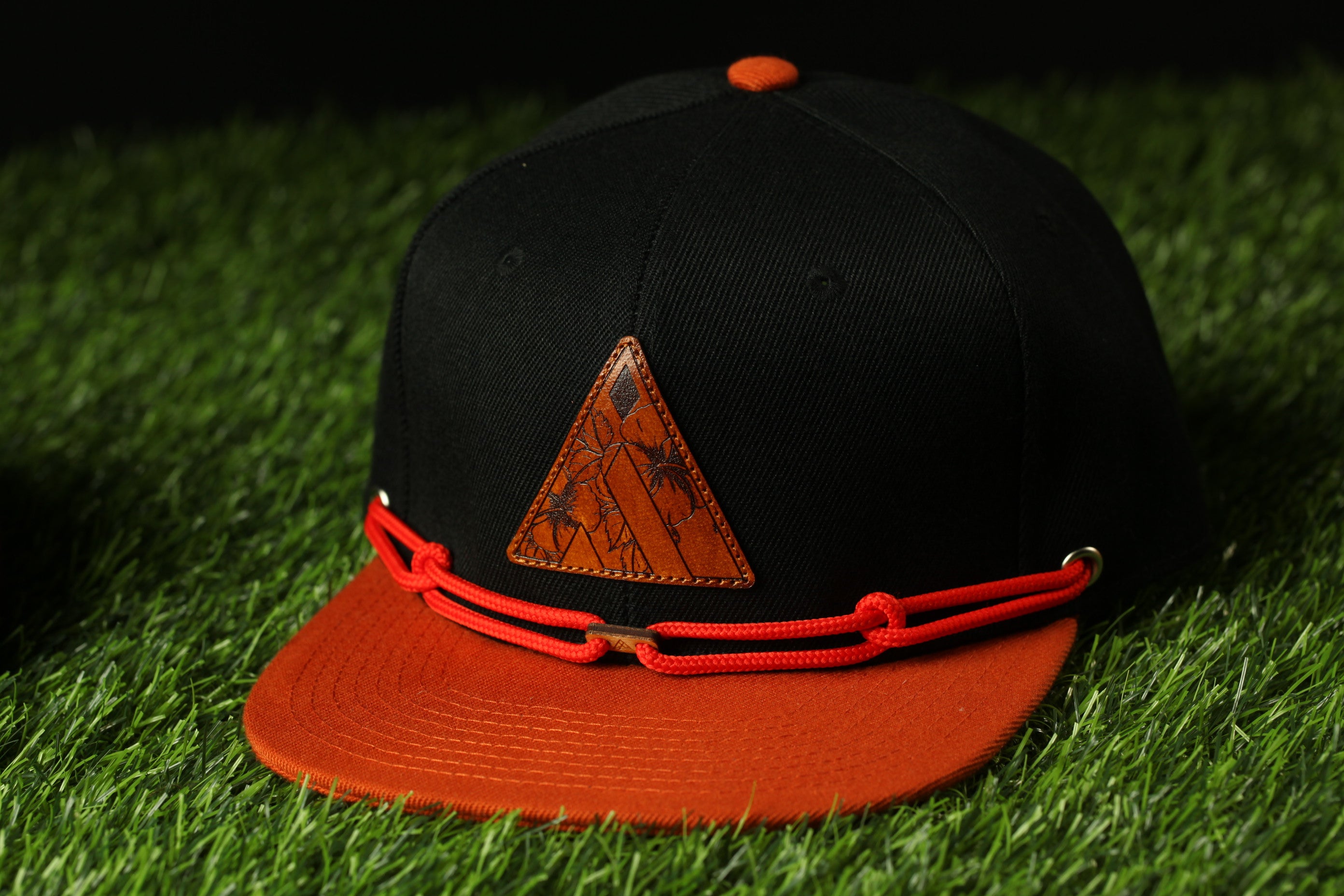 A black baseball cap with an orange band and a triangular patch on the front, placed on a grassy surface.