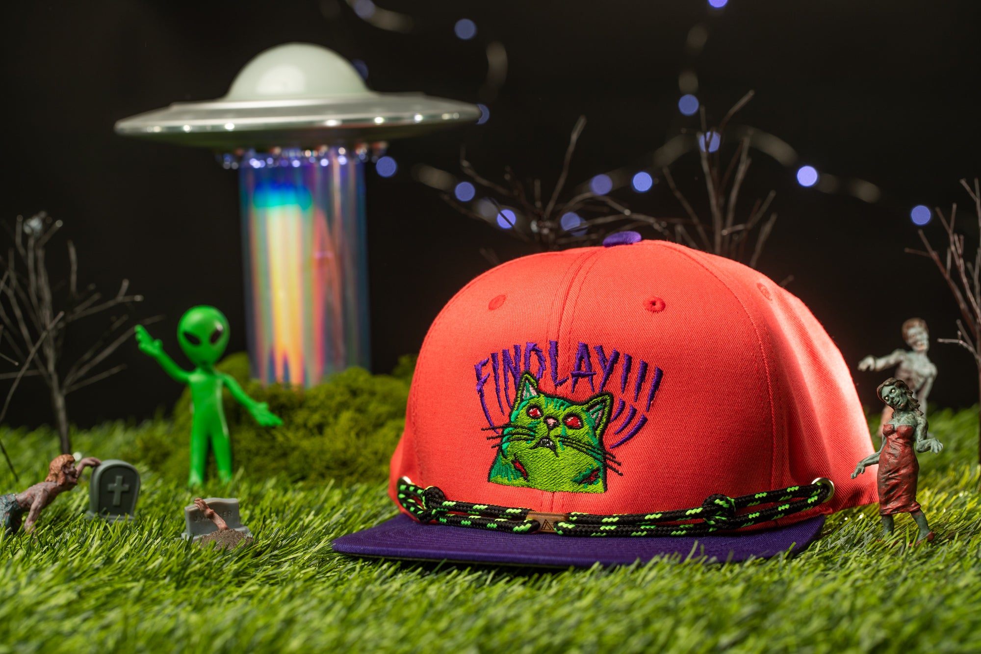 a red baseball cap with a cat design, surrounded by various toys and figurines, including a green alien figure, a zombie, and a spaceship.