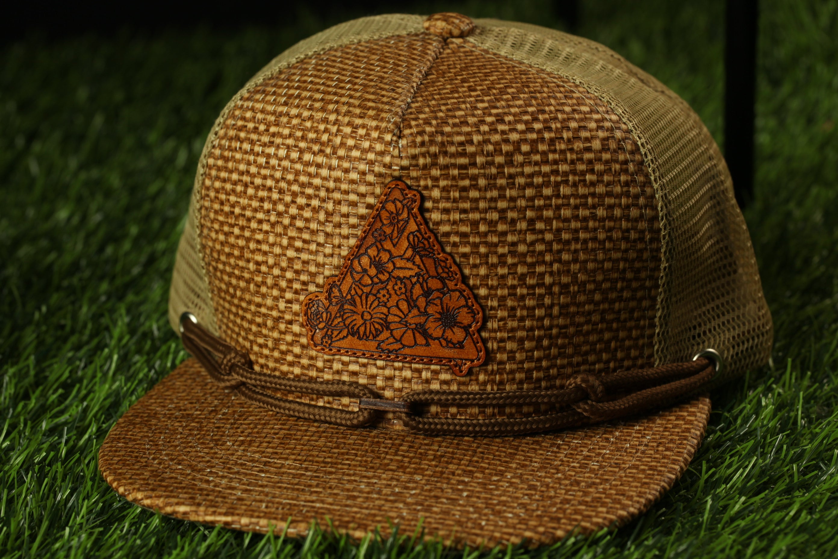 A brown woven baseball cap with a floral pattern on the front, placed on a grassy surface.