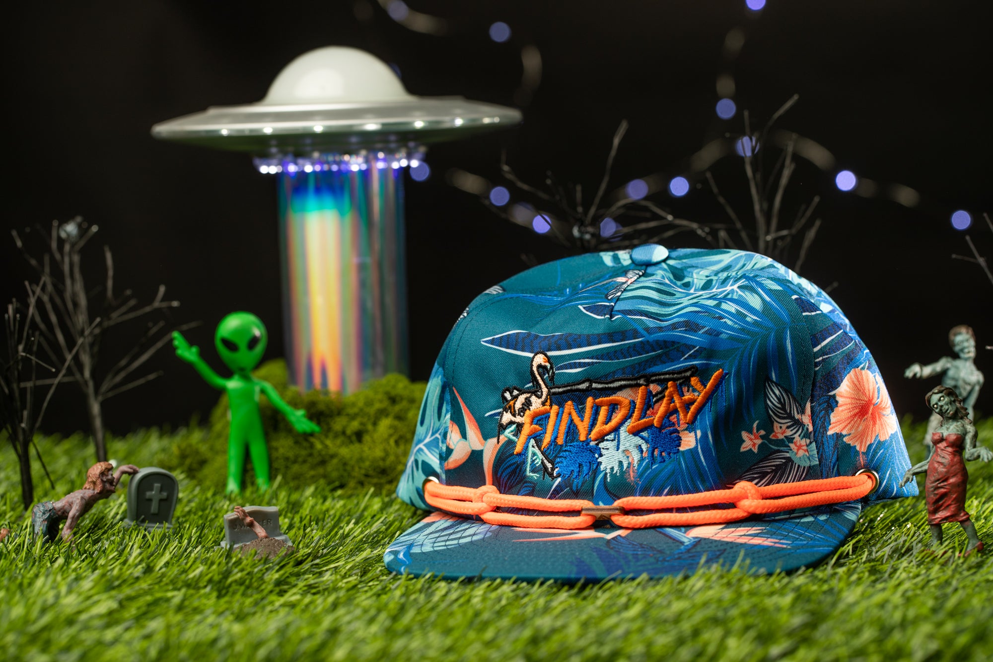 A colorful baseball cap with the word "Findlay" printed on it, surrounded by various small objects such as a green alien figurine, a skull, and a miniature alien spacecraft.