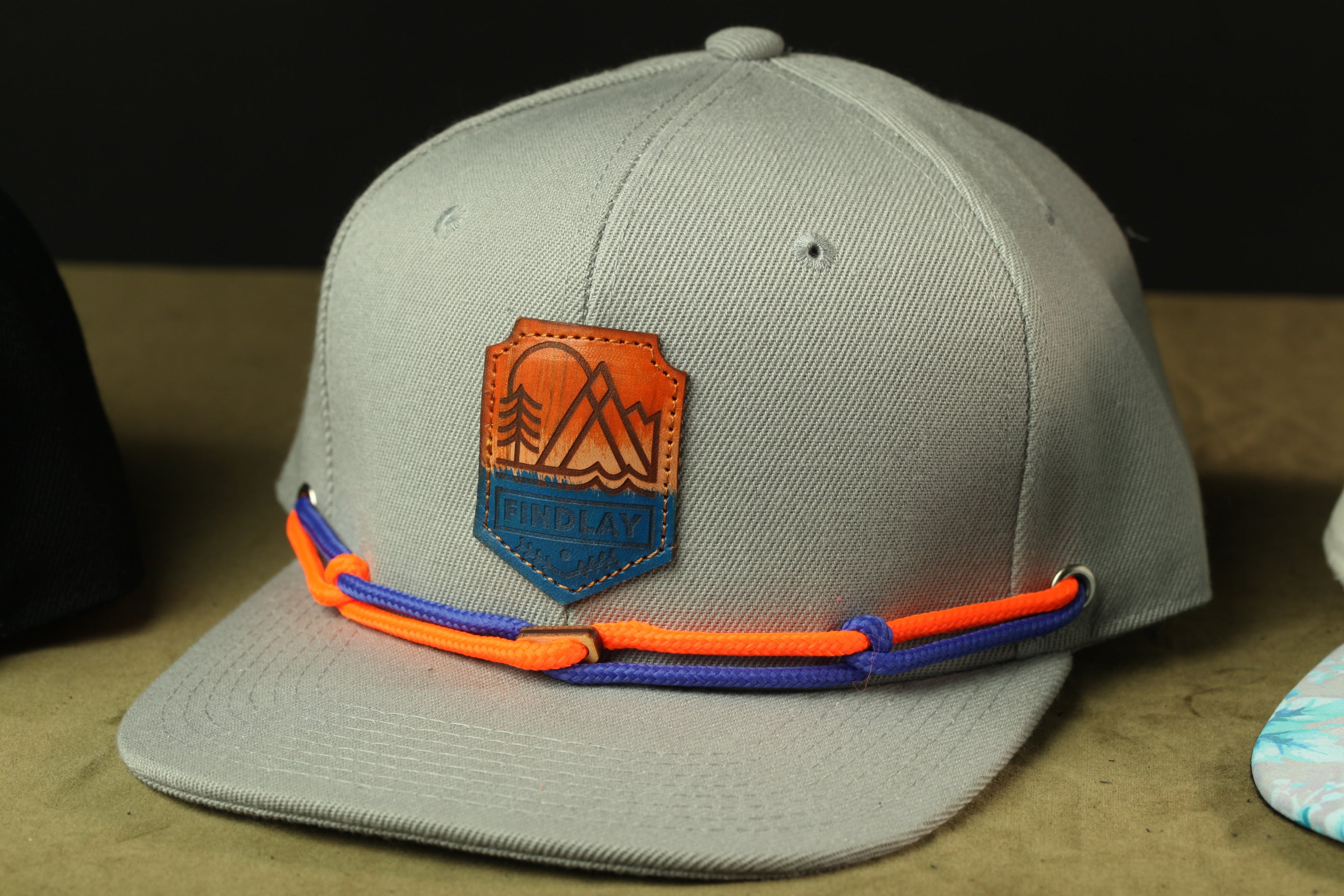 A gray baseball cap with a leather patch and orange and blue rope accents.