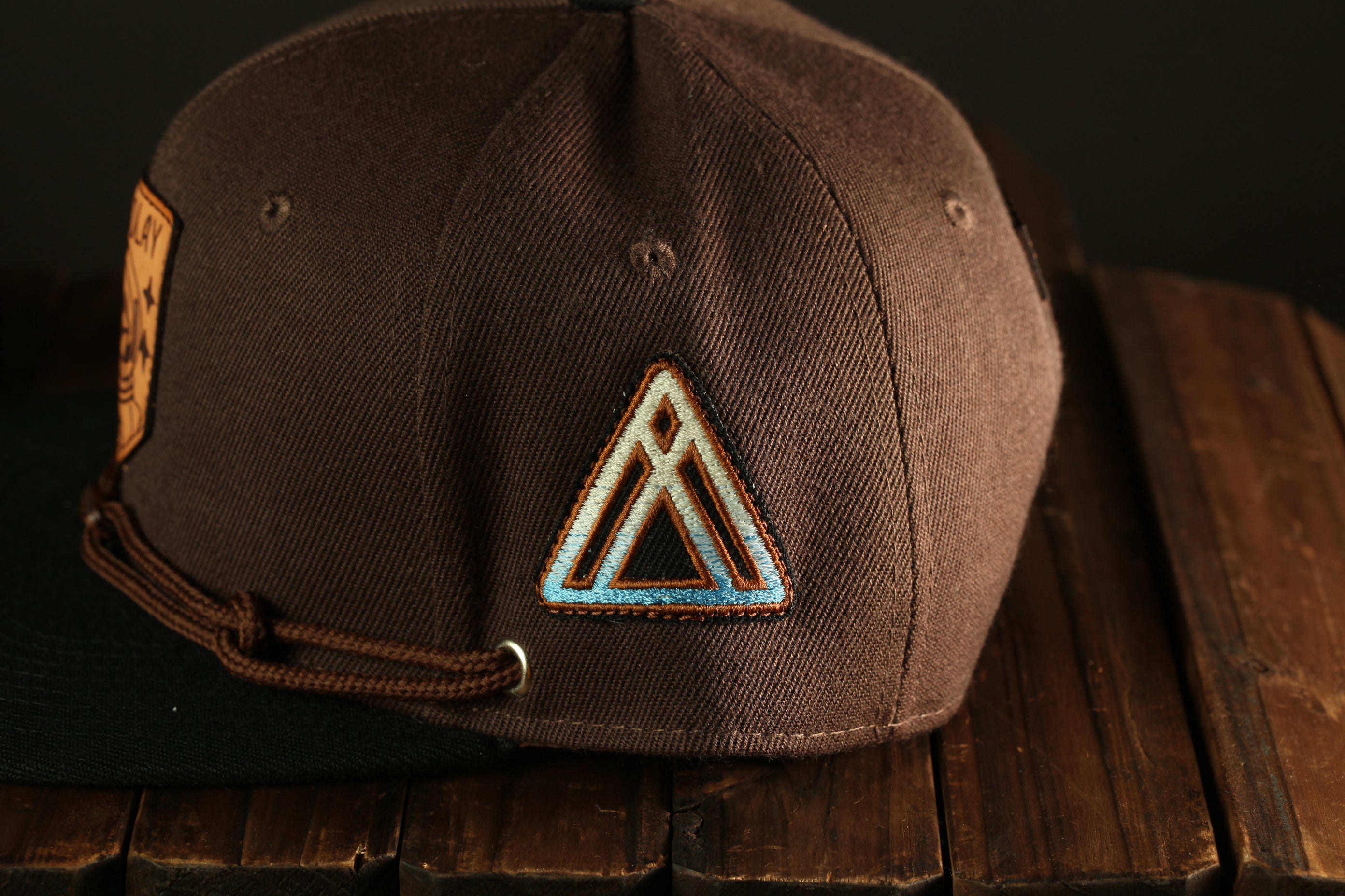 A brown baseball cap with a triangular patch on the front, resting on a wooden surface.