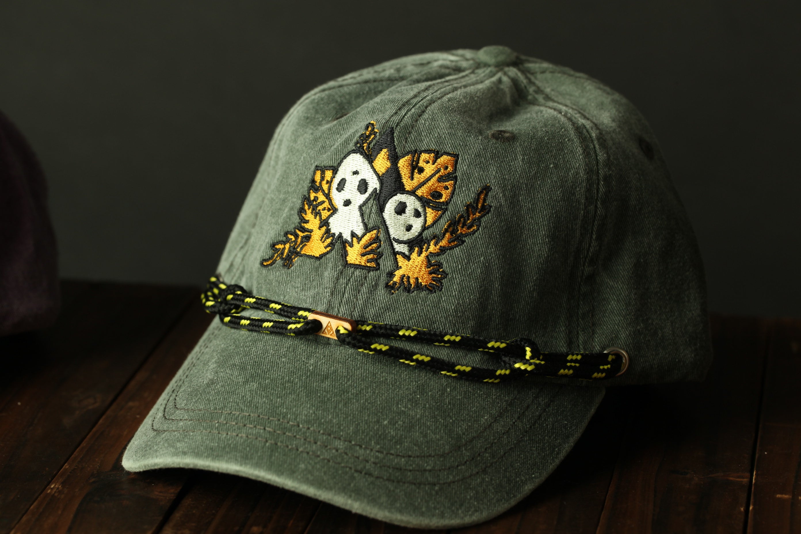 A gray baseball cap with a black and yellow embroidered design on the front, featuring two cartoon-like faces and leaves.