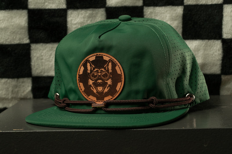 A green baseball cap with a brown leather patch featuring a cartoon dog wearing goggles.