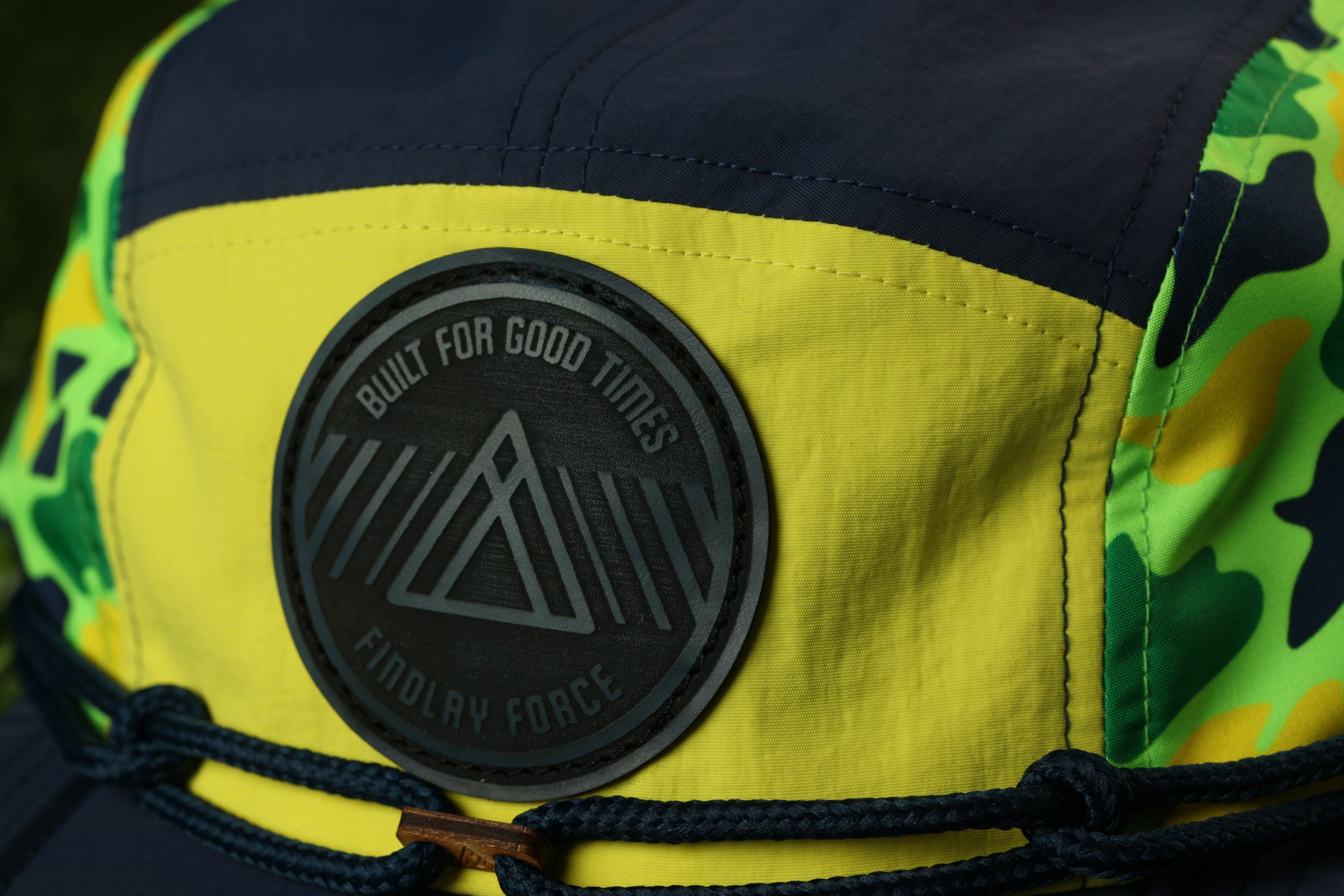 A close-up view of a yellow and black cap with a black circular patch on the front, featuring a graphic design and text.
