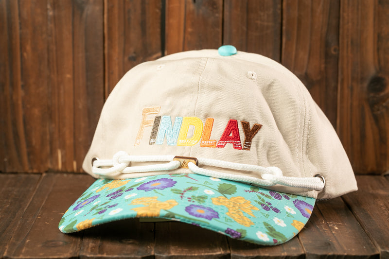 A beige baseball cap with a floral pattern and the word "FINDlay" embroidered on it, placed on a wooden surface.