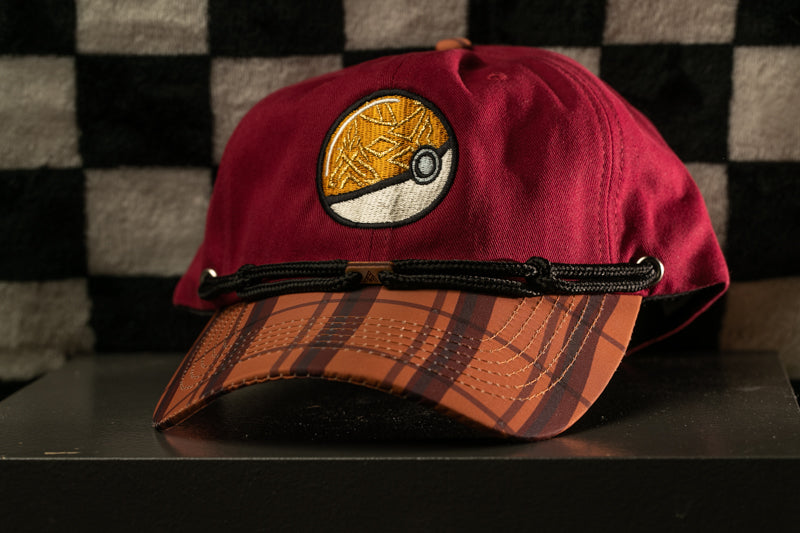A maroon baseball cap with a Pokemon-inspired design, featuring a circular patch with a yellow and orange Pokemon symbol, is displayed on a black surface against a checkered background.