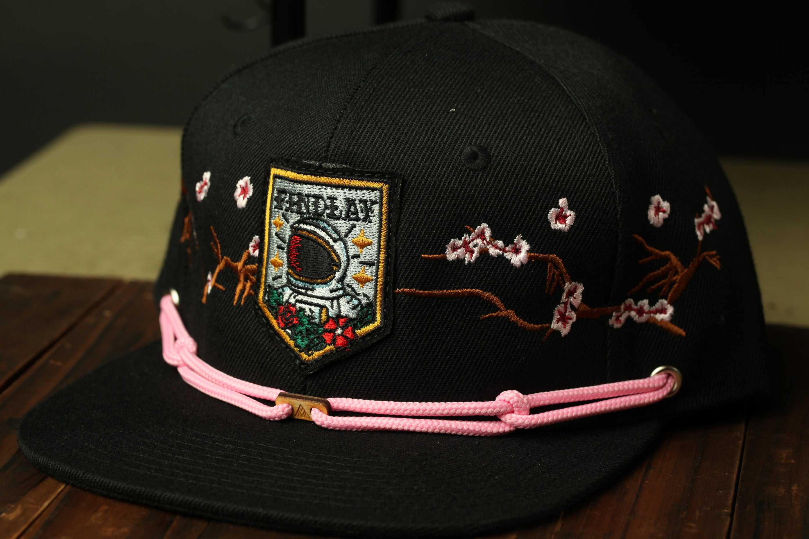 A black baseball cap with a pink braided strap and a patch featuring a space helmet and cherry blossom flowers.