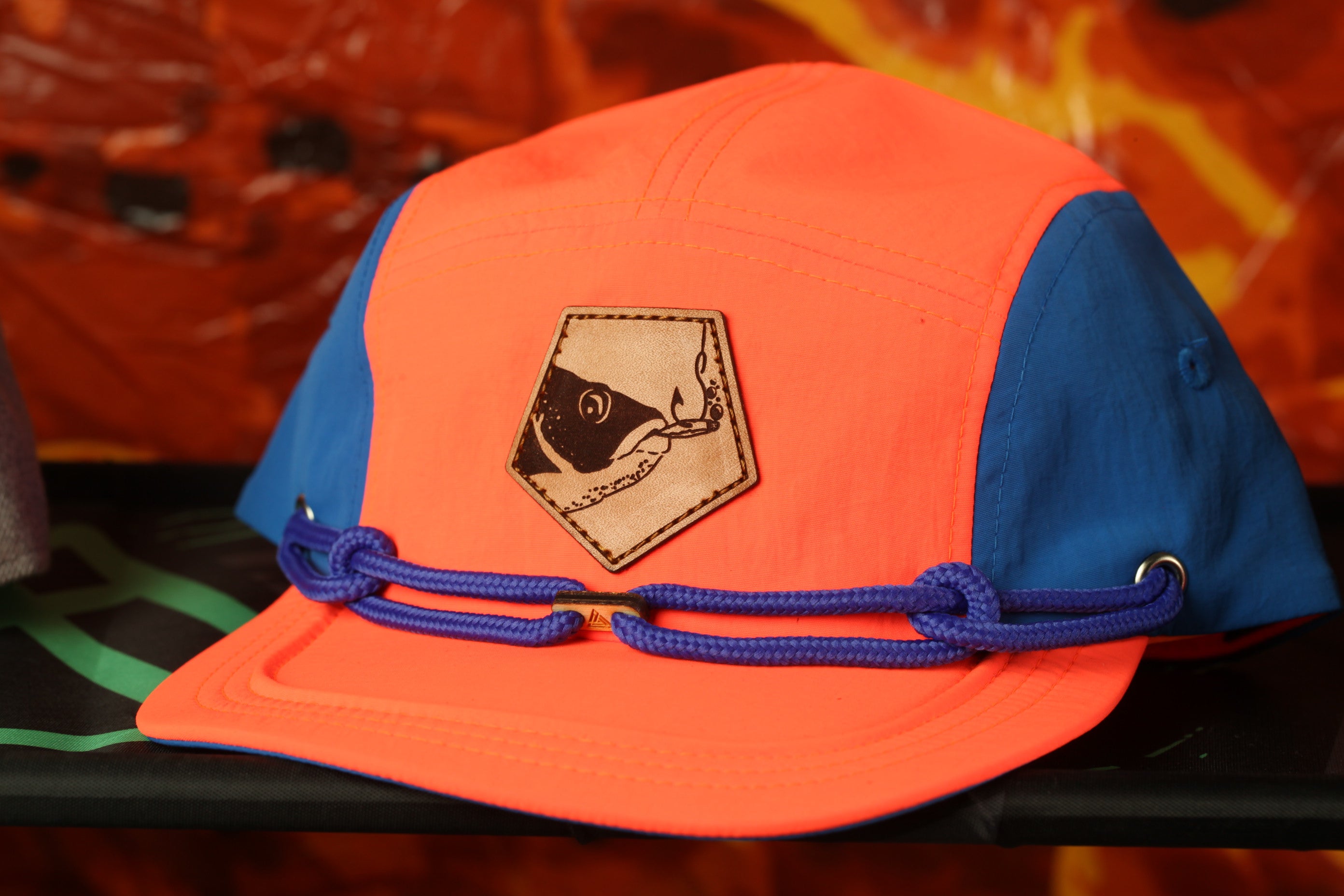 An orange and blue baseball cap with a leather patch featuring a fish design, resting on a black surface.