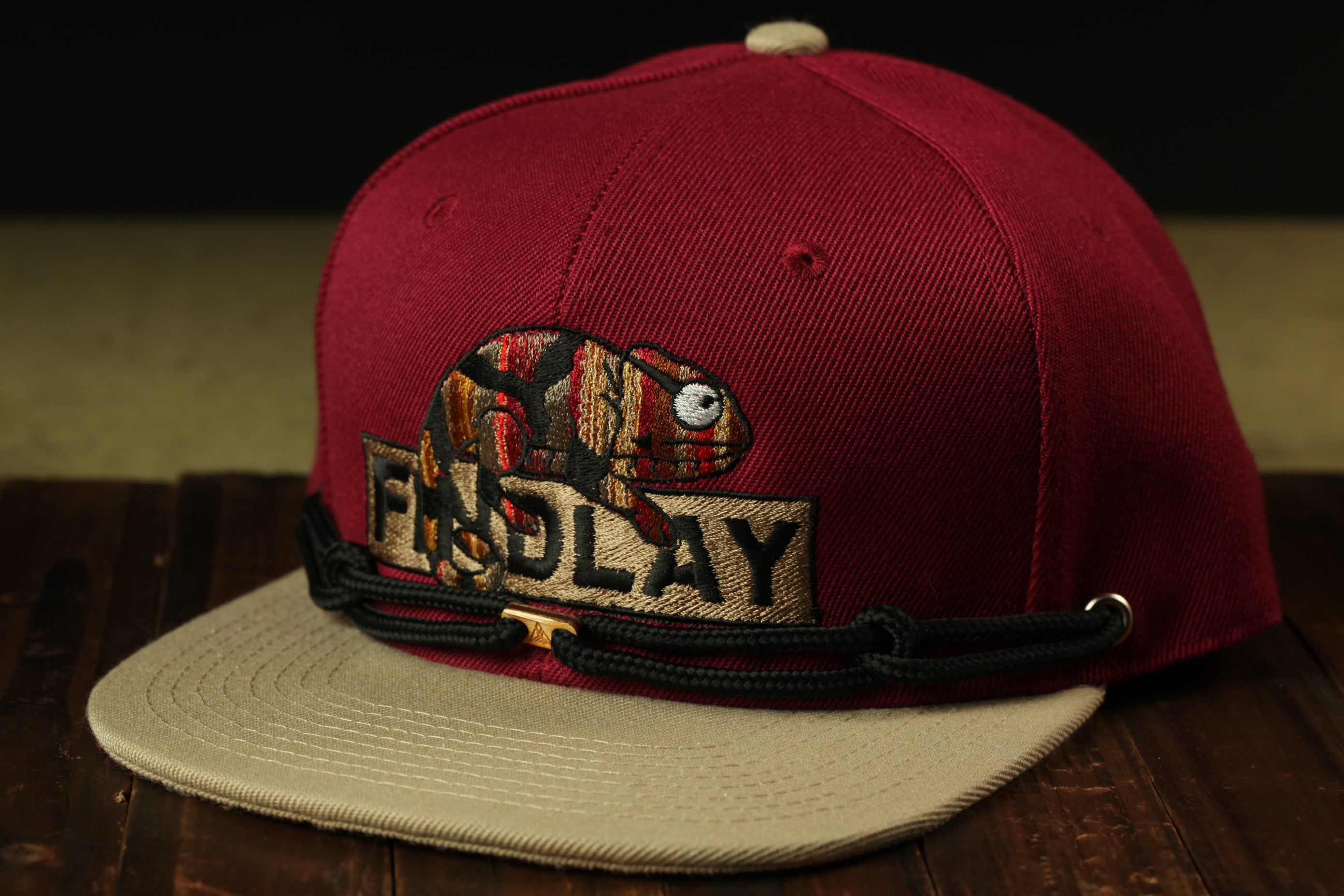 A maroon baseball cap with a graphic of a chameleon and the word "FINDlay" printed on it, sitting on a wooden surface.
