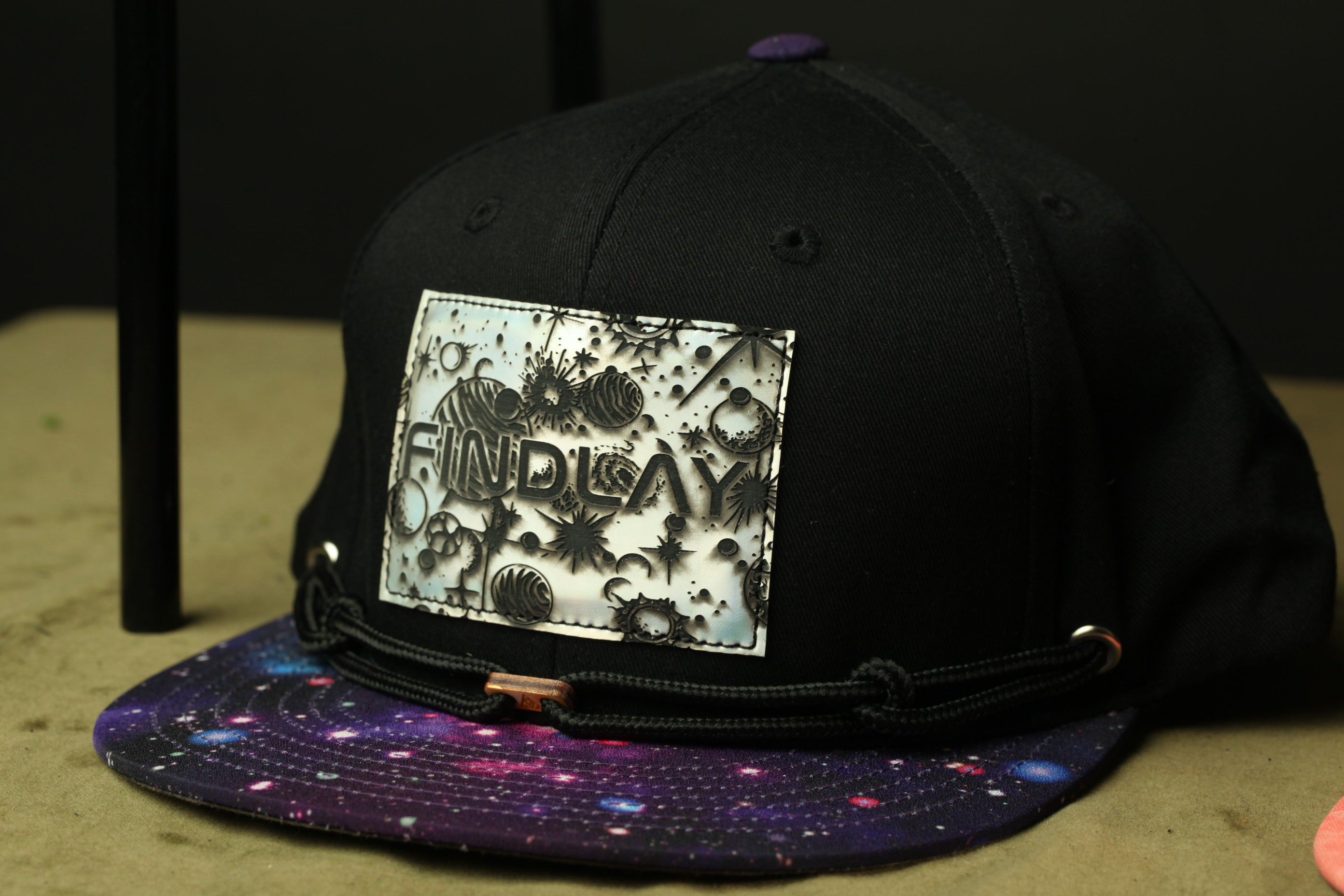 A black baseball cap with a galaxy print pattern and a patch on the front displaying the word "FIND" in a stylized font.