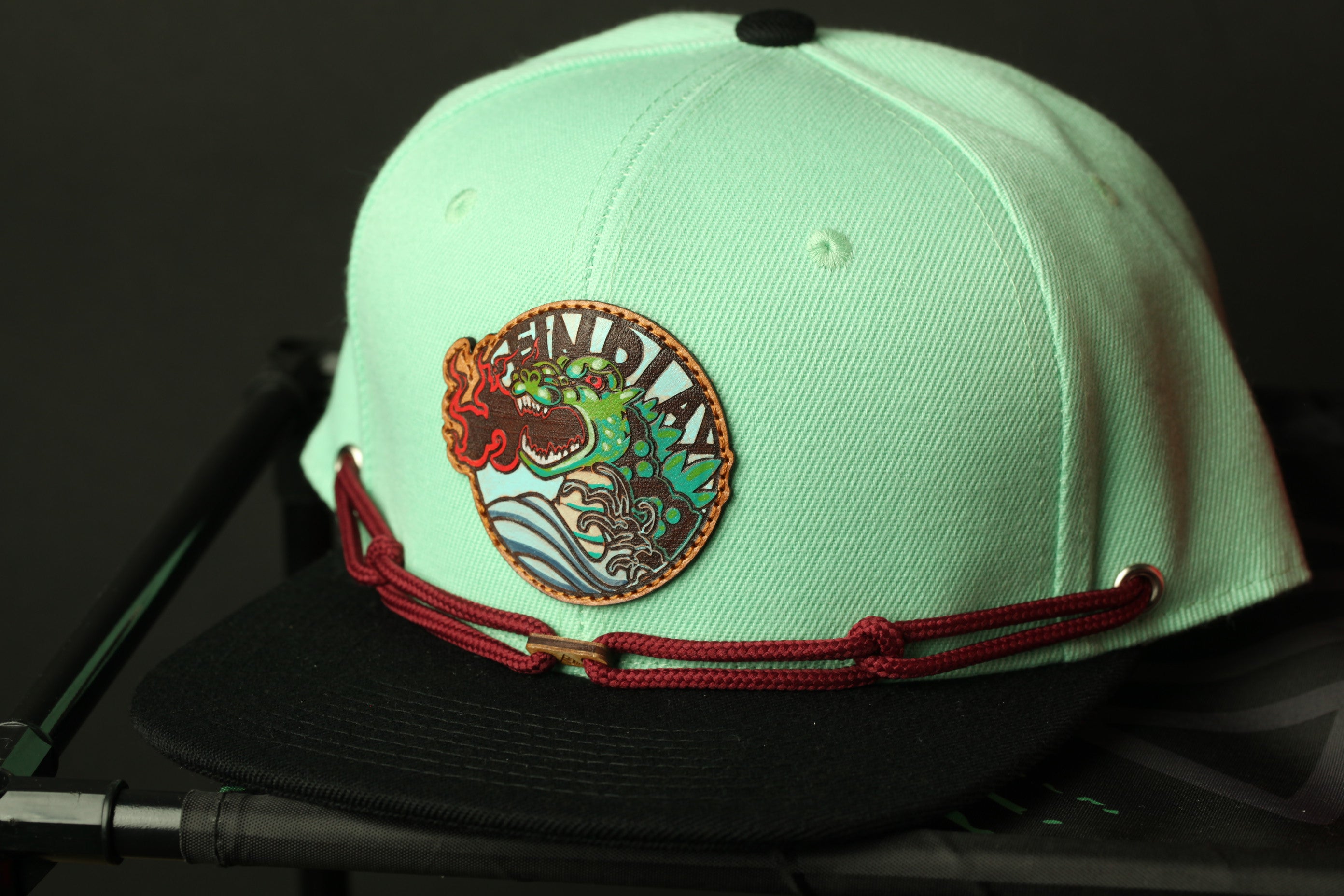 A mint green baseball cap with a red and black adjustable strap and a circular patch on the front.