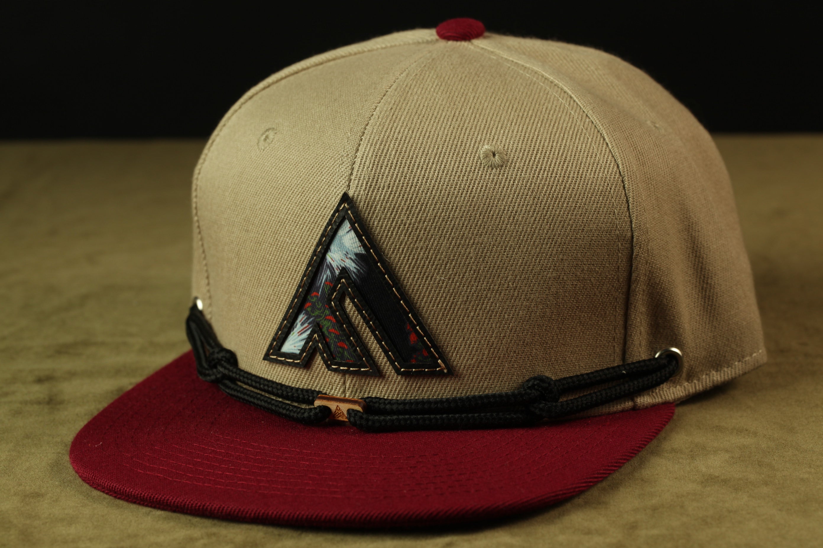 A tan baseball cap with a red brim and a black and red logo on the front, placed on a brown surface.
