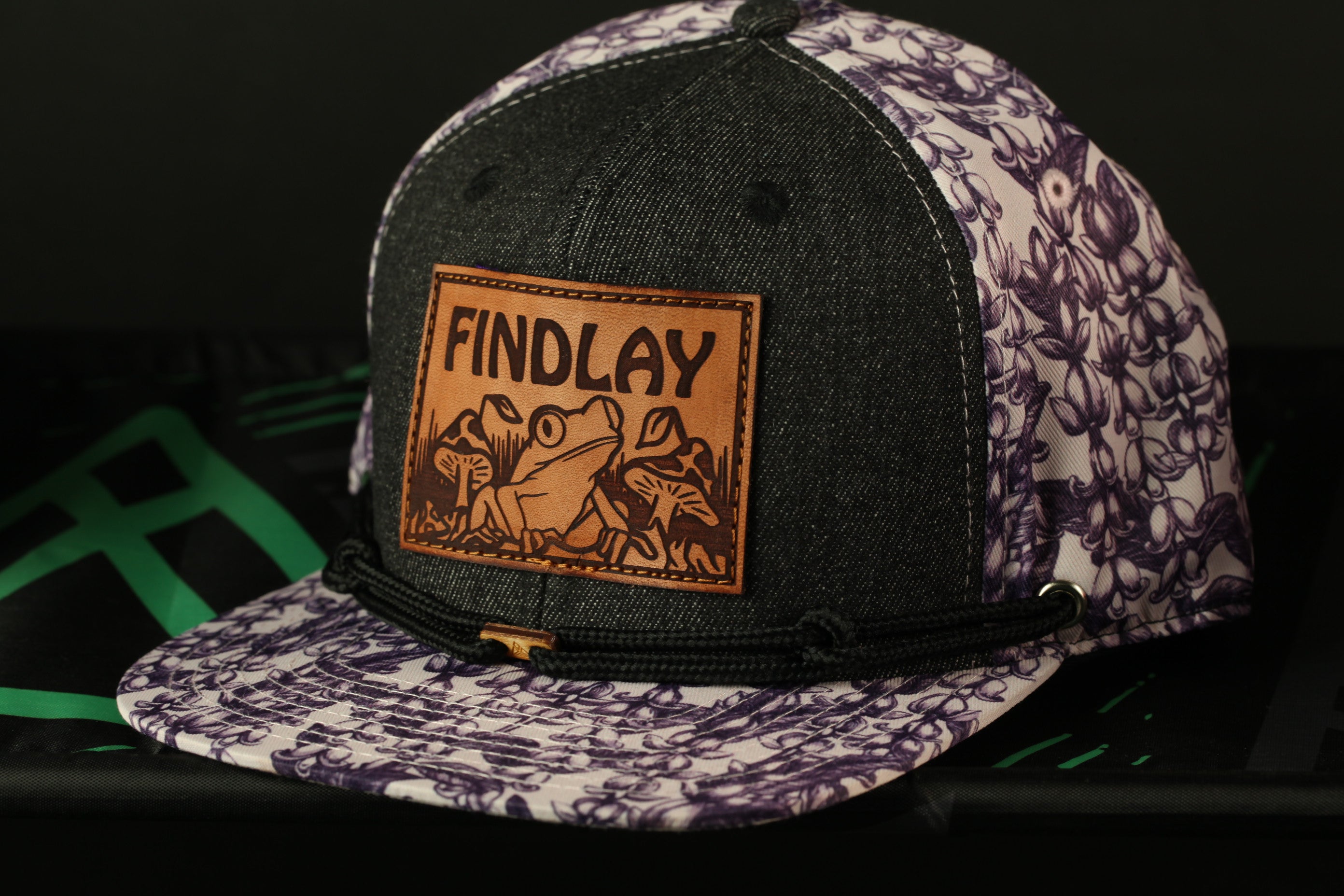 A black and purple baseball cap with a leather patch on the front, featuring a frog and the word "FINDLAY" printed on it.