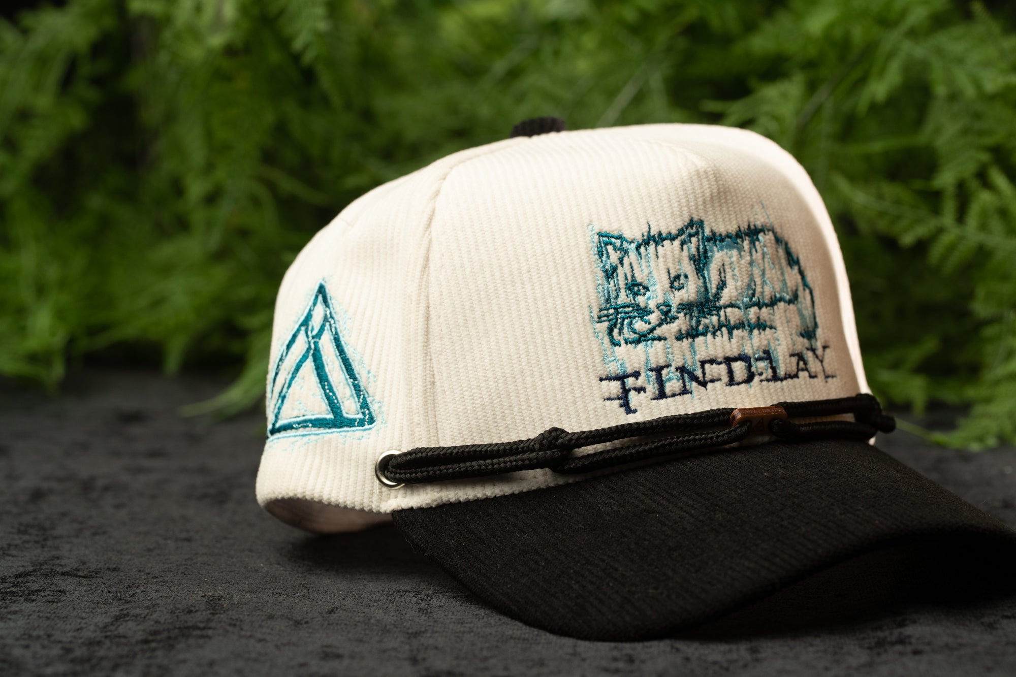 A white baseball cap with a black brim, featuring a cat embroidered on the front, is displayed against a blurred green background.