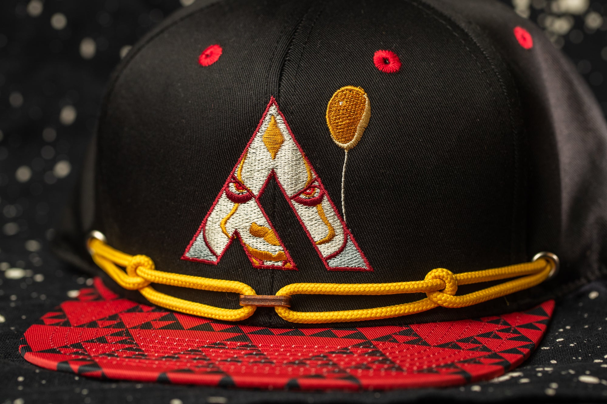 A close-up image of a black baseball cap with a red and yellow patterned brim, a gold chain strap, and a red and gold logo on the front.