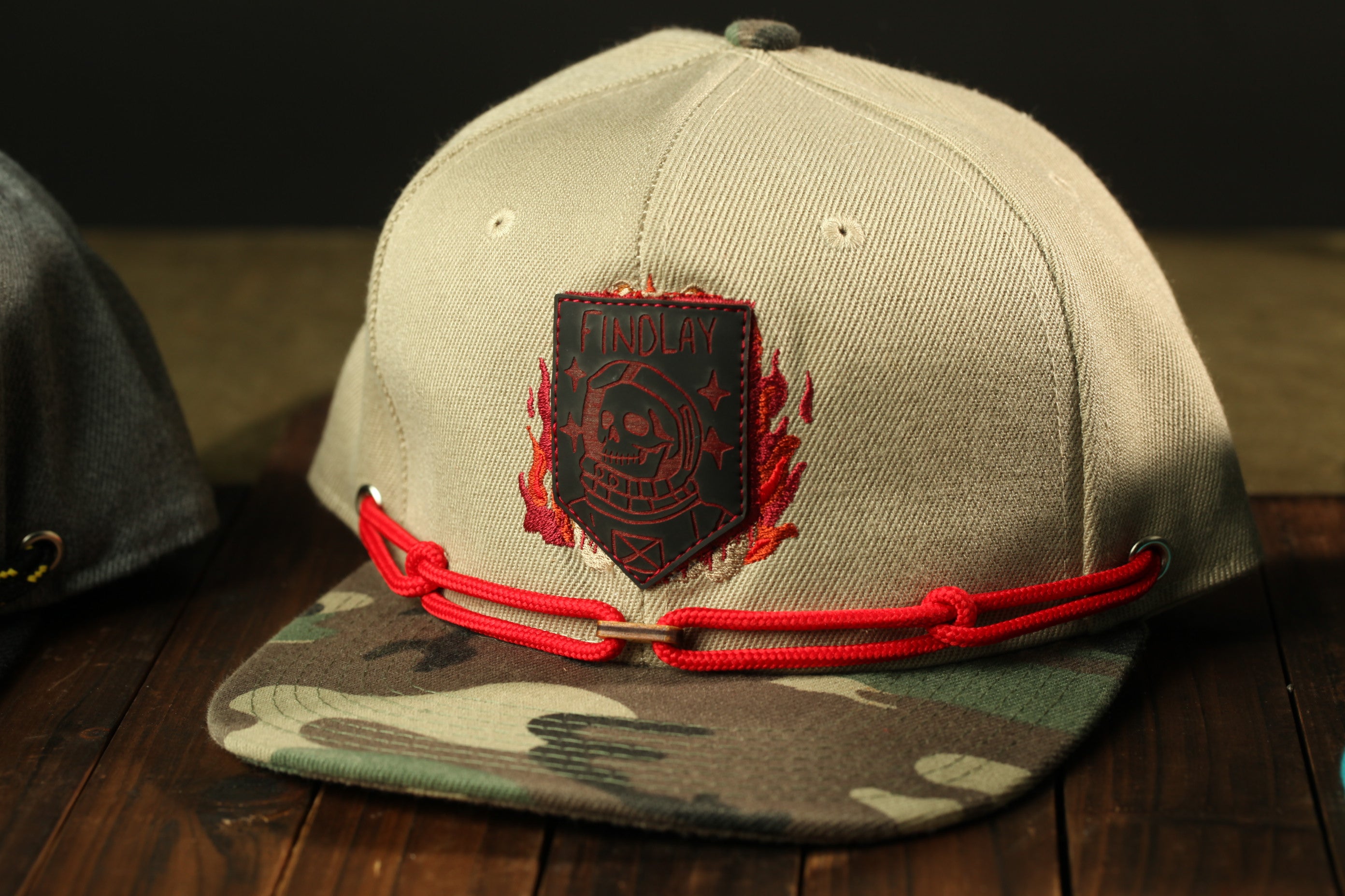 A beige baseball cap with a red chain and a black patch featuring a skull and crossbones design, placed on a wooden surface.