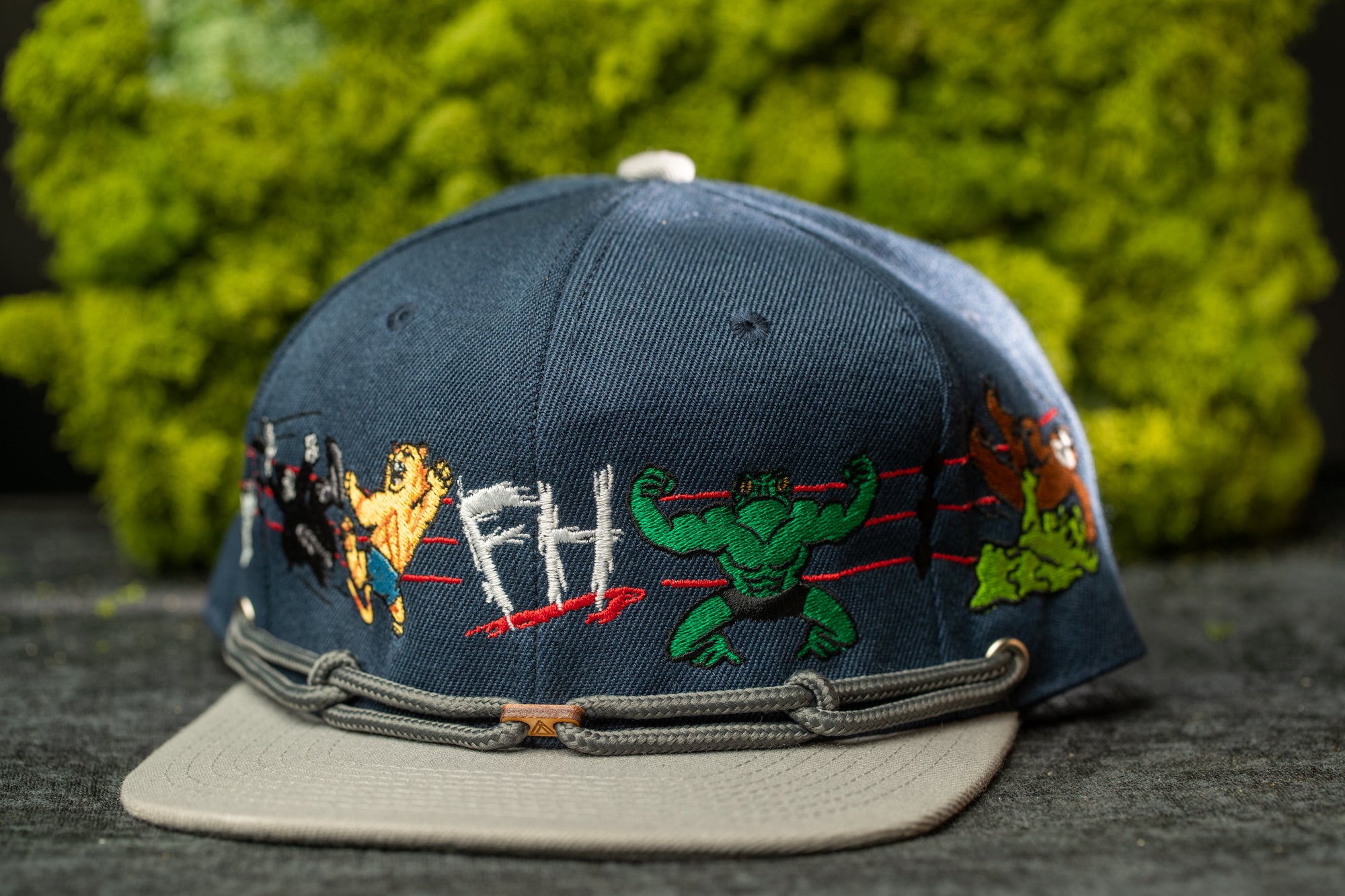 A blue baseball cap with various cartoon characters and text embroidered on it, placed on a surface with a green mossy background.