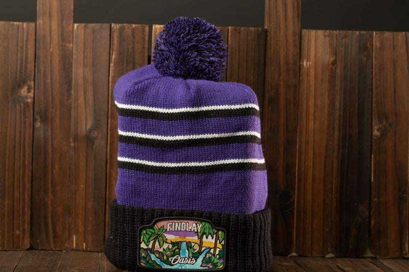 A purple and black striped beanie with a pom pom on top, placed on a wooden surface against a wooden background.