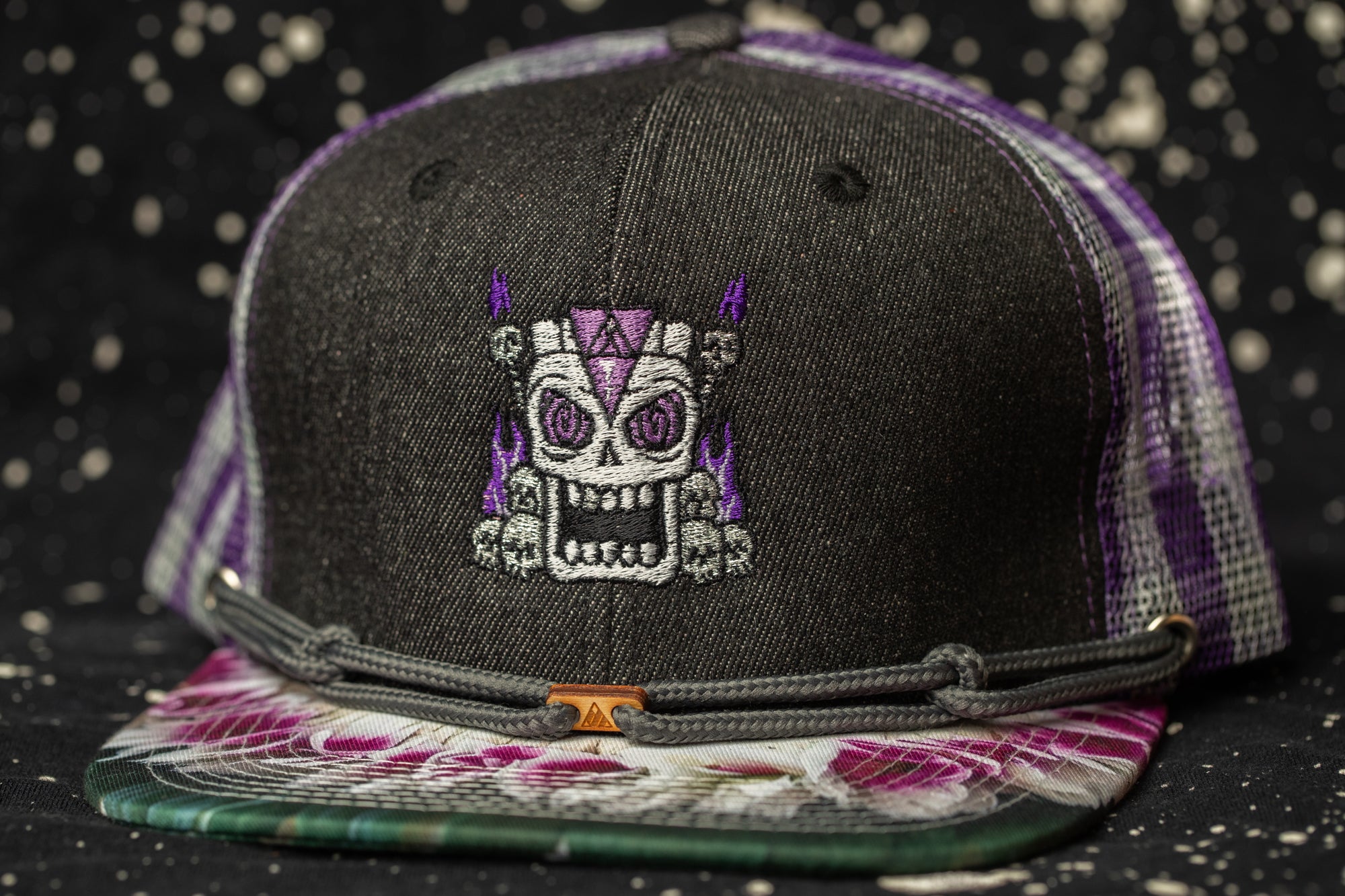 A black baseball cap with a purple and white patterned brim, featuring an embroidered skull design on the front.