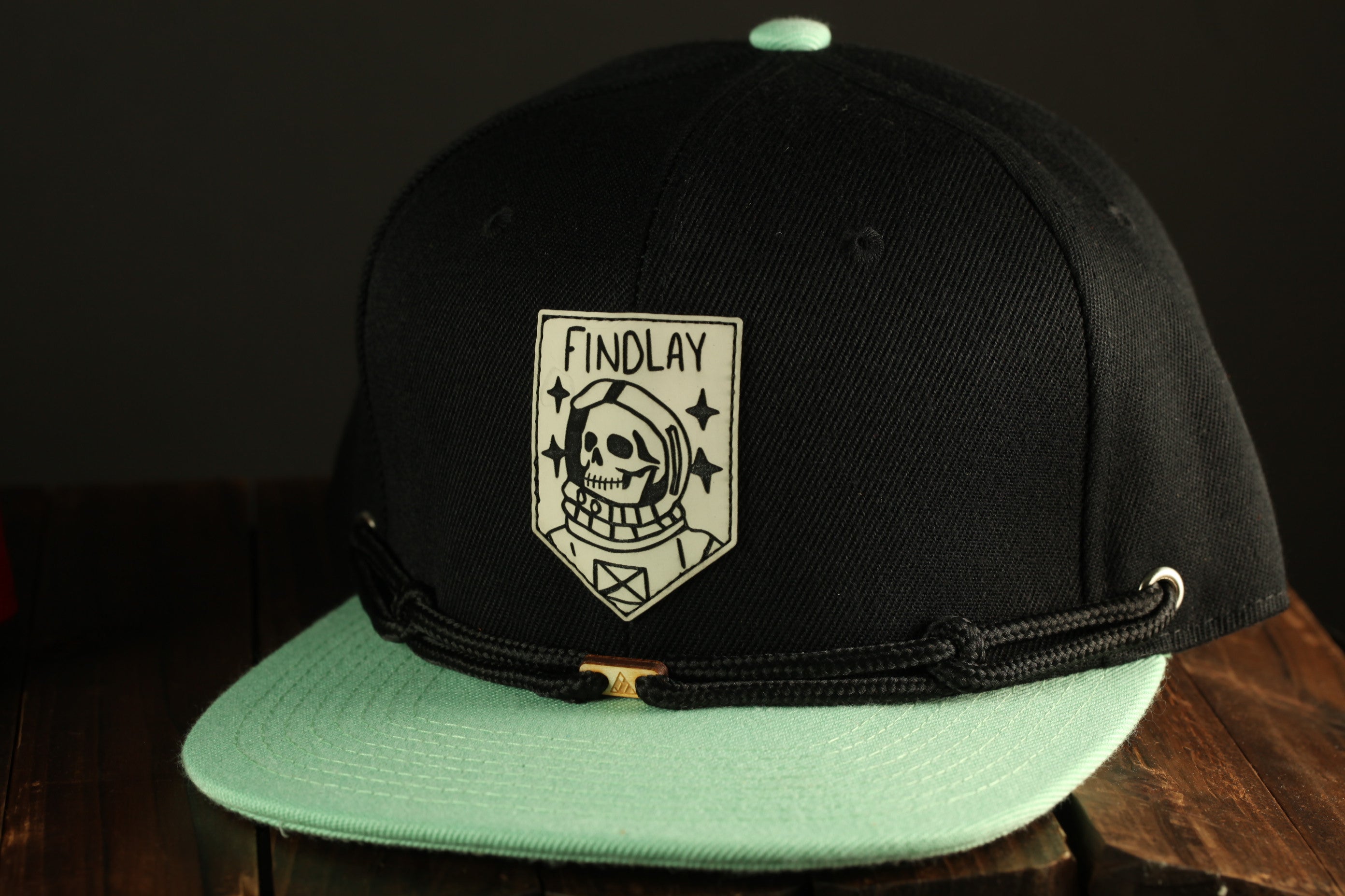 A black baseball cap with a teal brim and a white skull and crossbones logo on the front, placed on a wooden surface.
