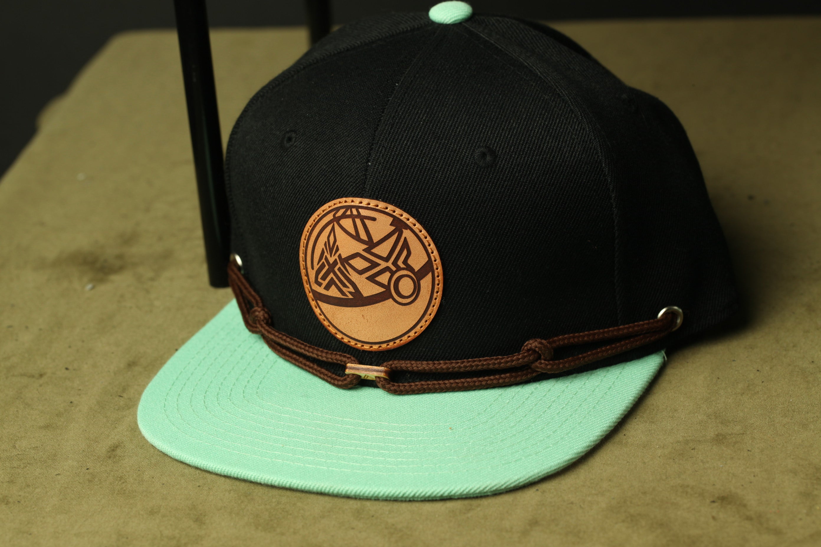 A black baseball cap with a green brim and a brown leather patch on the front, placed on a wooden surface.